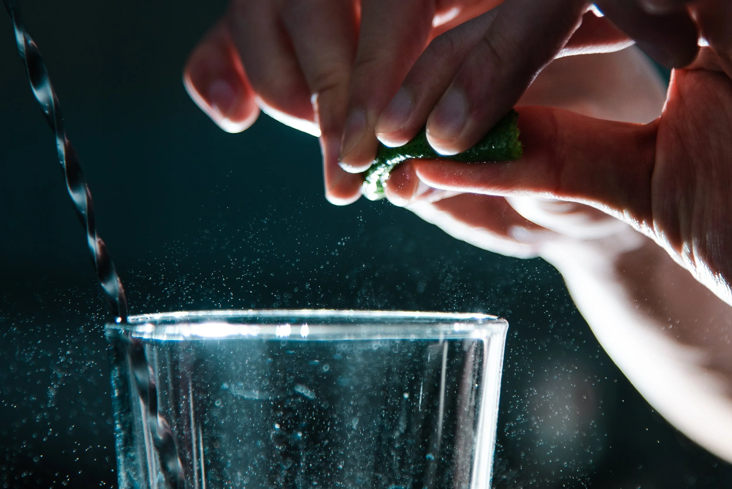 Close-up of hands squeezing a fresh lime over a glass of water, with droplets of lime juice falling into the water.