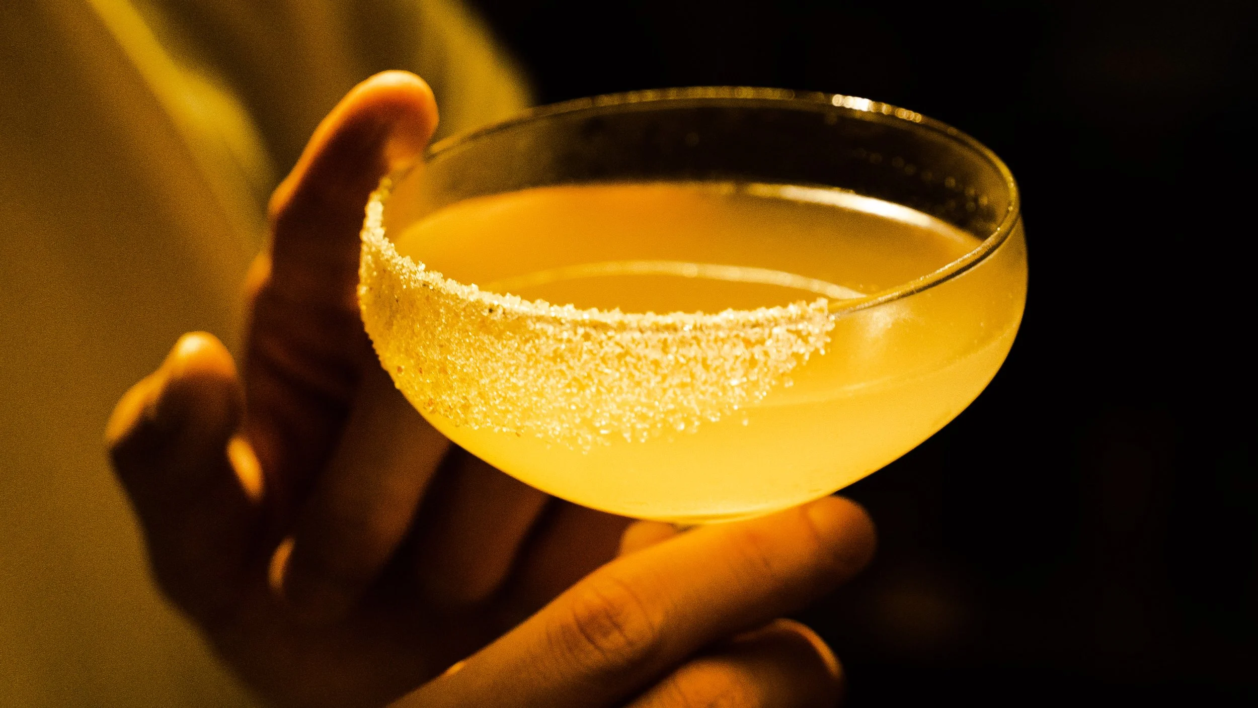 A yellow cocktail in a coupe glass with a sugar-rimmed edge, held by a hand against a dark background.