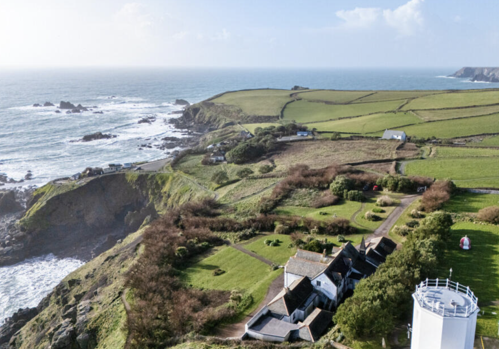 Coastal landscape with green fields, houses, and a white lighthouse near the ocean and cliffs.