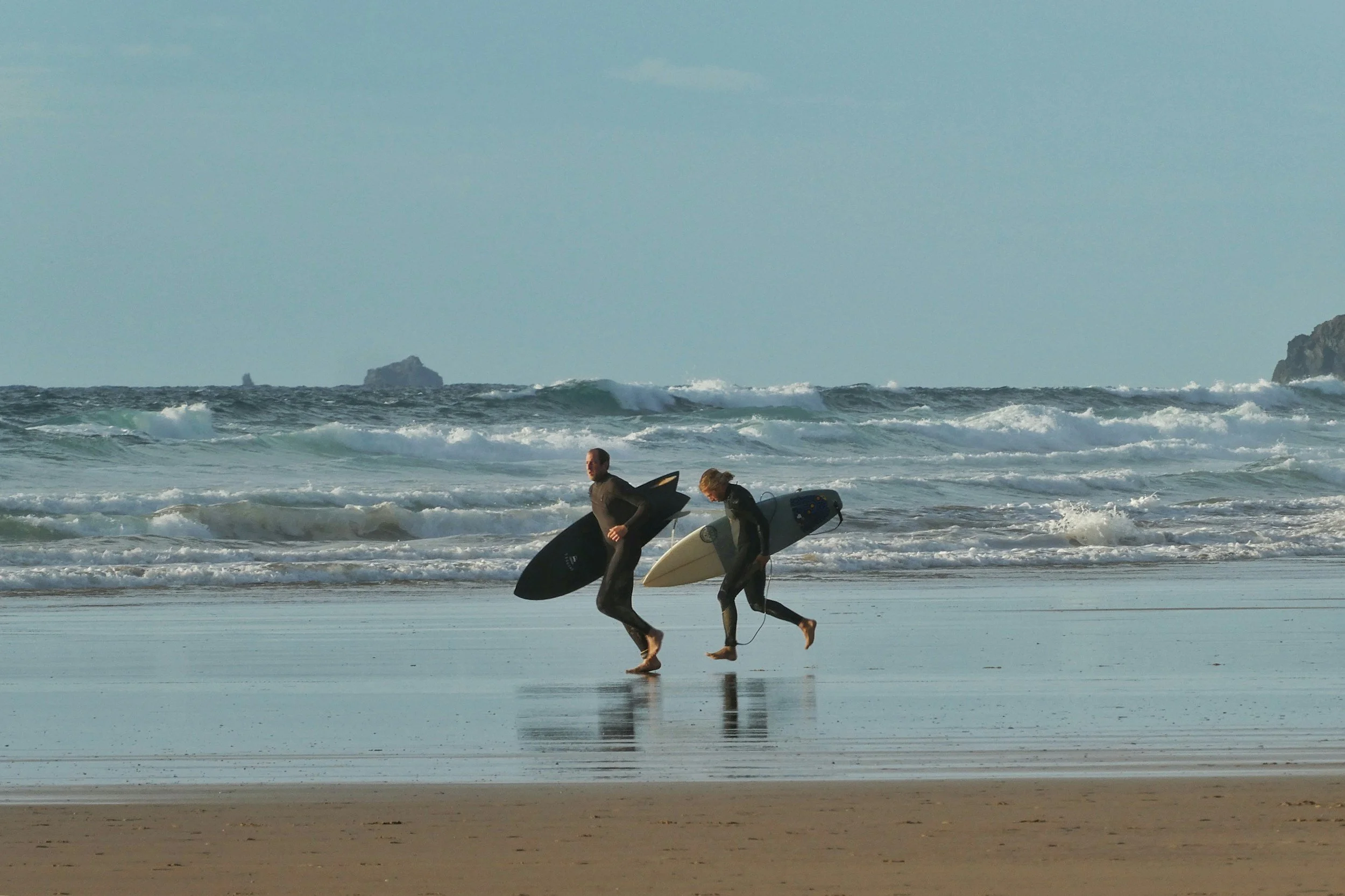 Two surfers in wetsuits walking along the beach carrying surfboards with ocean waves and distant rocks in the background.