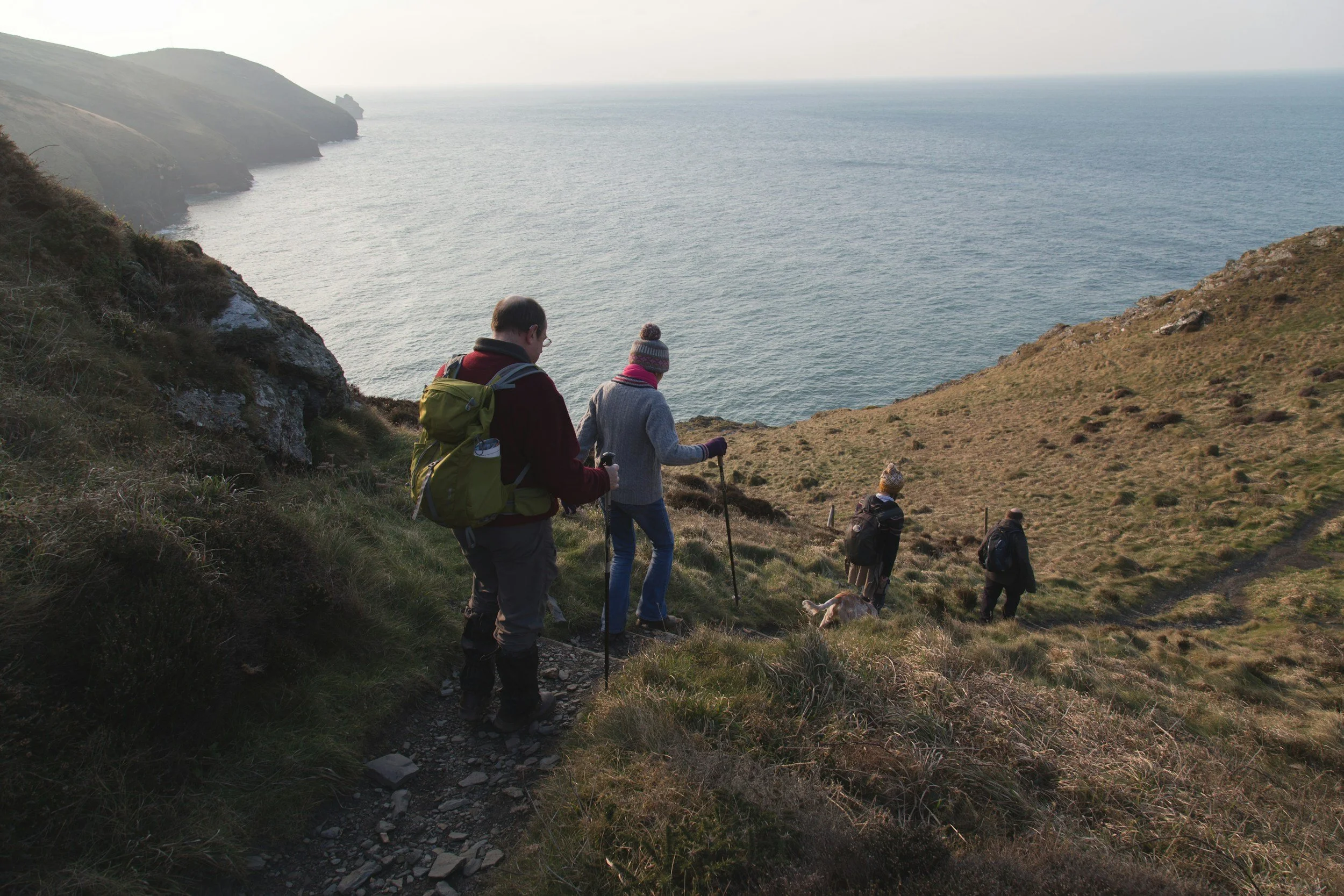Group of hikers walking down a grassy trail along a coastal hillside with cliffs and the ocean in the background.