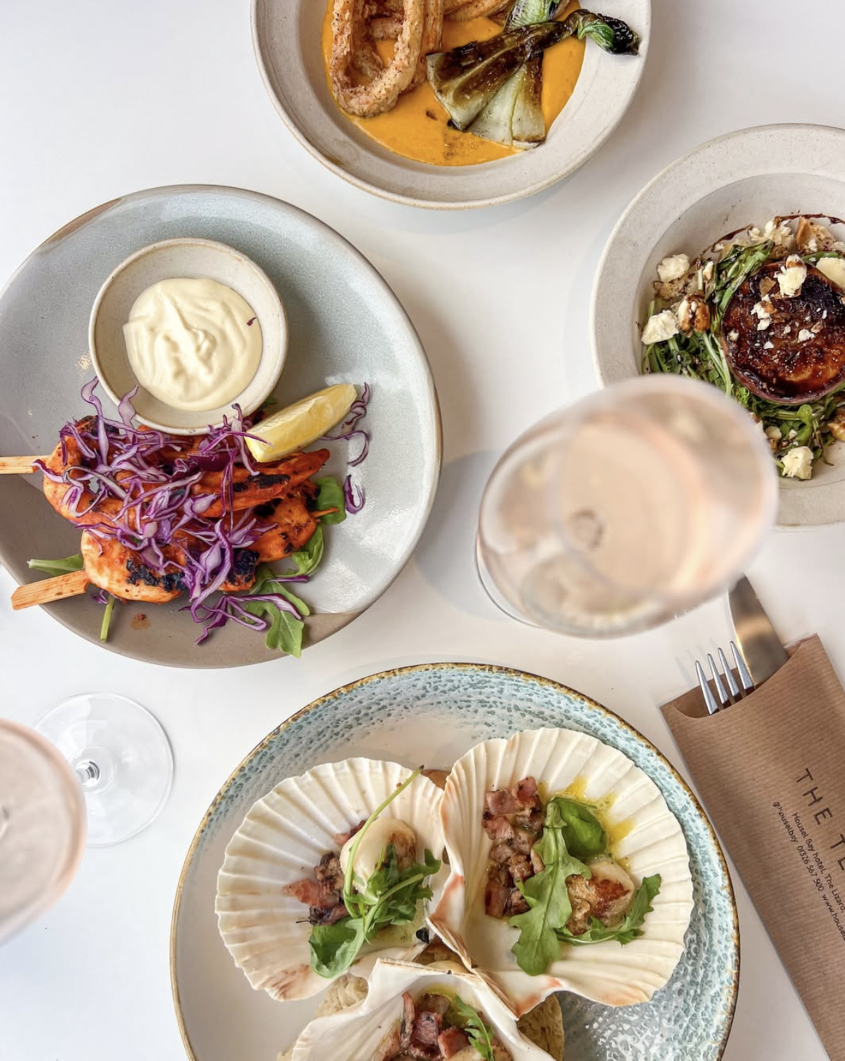 Assorted dishes including grilled chicken skewers with purple cabbage and lemon, roasted vegetables in a creamy sauce, glazed meatball with greens, and seafood in shells with greens, along with glasses of wine on a white table.