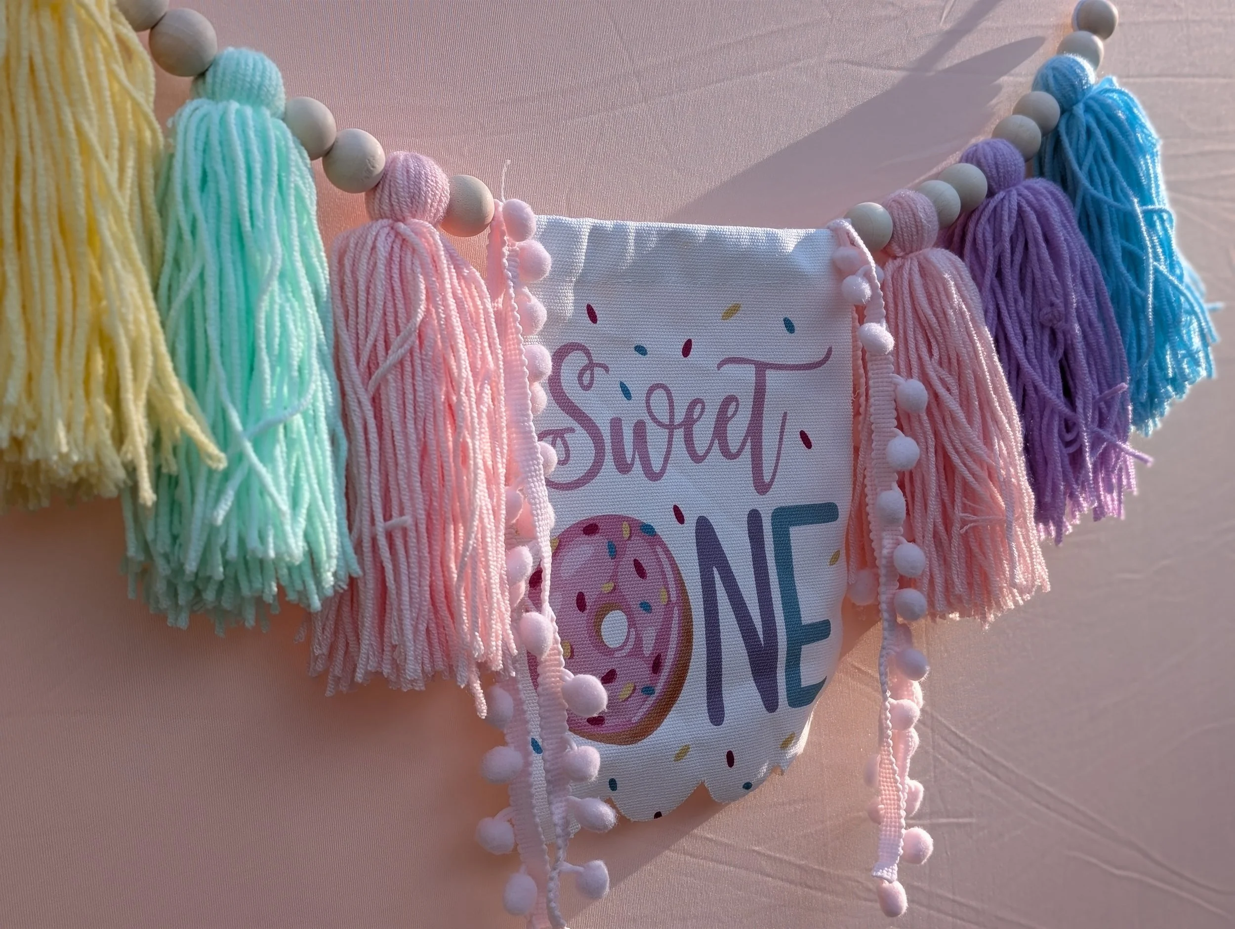 Colorful pastel tassels hanging on a string with a white fabric banner that has the word "Sweet" and a donut illustration.