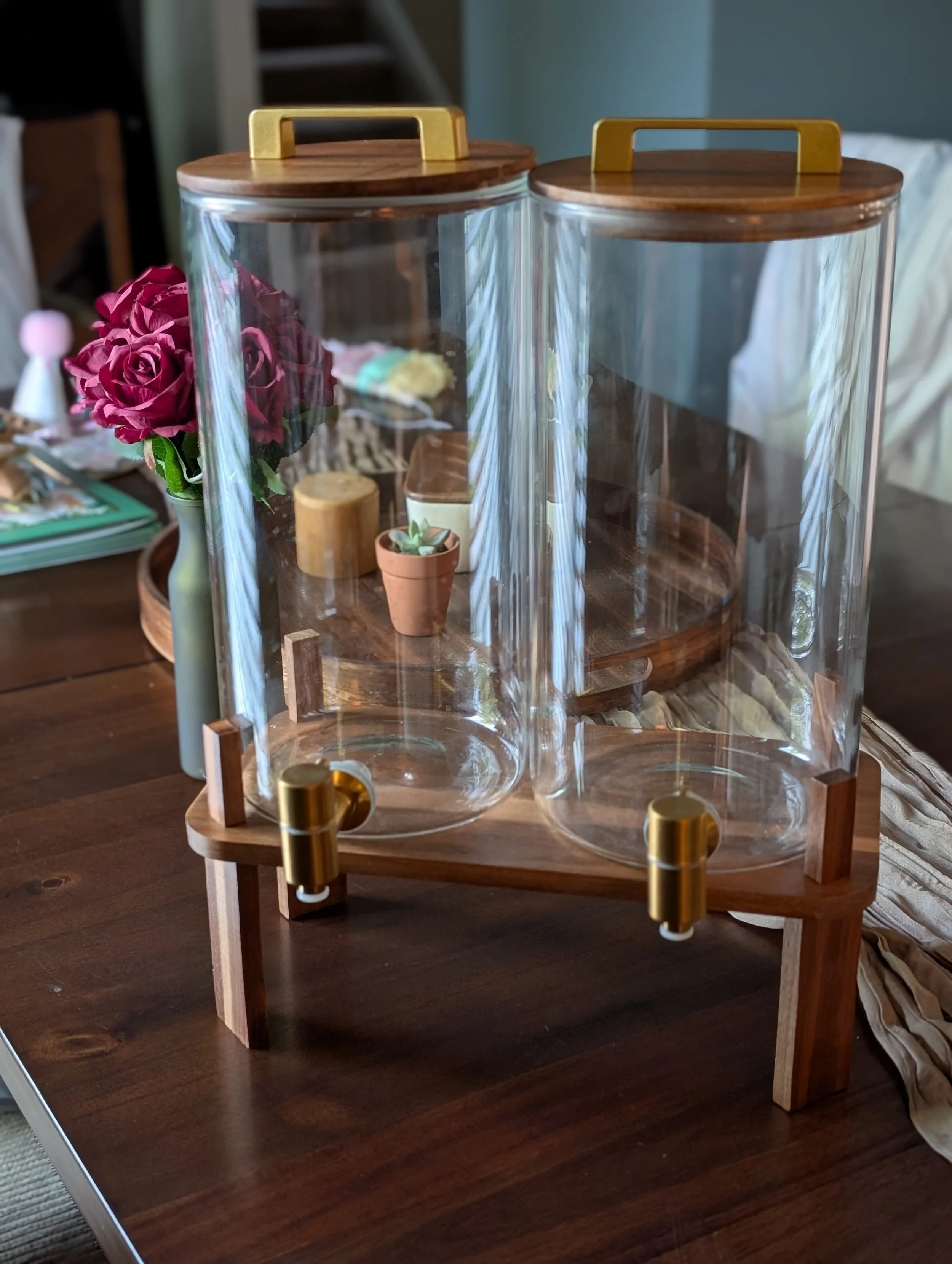 A wooden and glass display case with a wooden base and clear glass panels, sitting on a wooden table. Behind the display case, there are decorative items including a potted succulent, a small flower arrangement, and other assorted objects. Part of a 