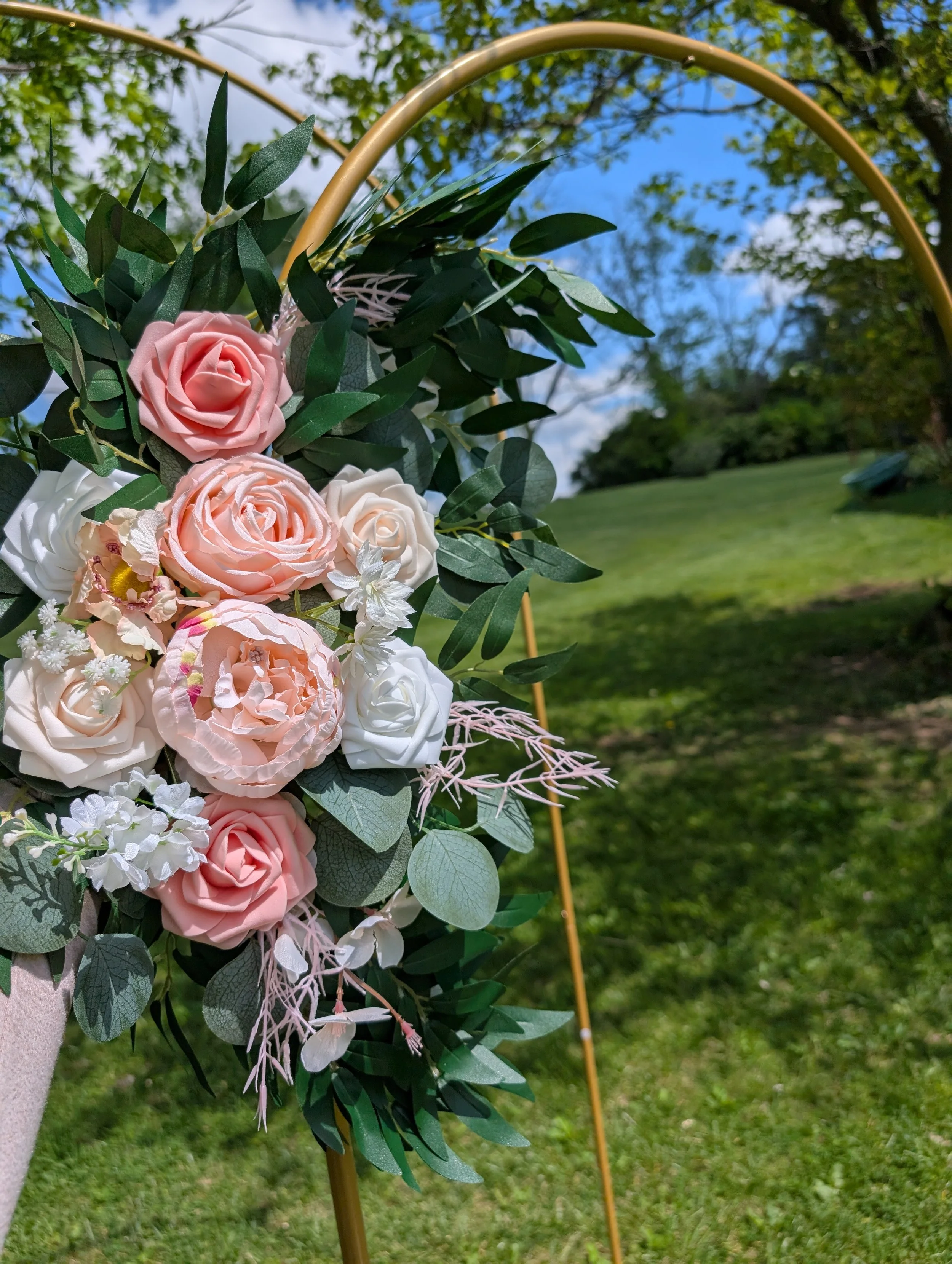 Close-up of a floral arrangement with pink, white, and cream roses, surrounded by green leaves, set outdoors with a tree and blue sky in the background.