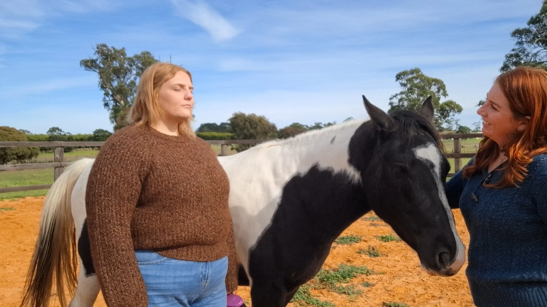 Two women and a black and white horse in a paddock, with a blue sky and green trees