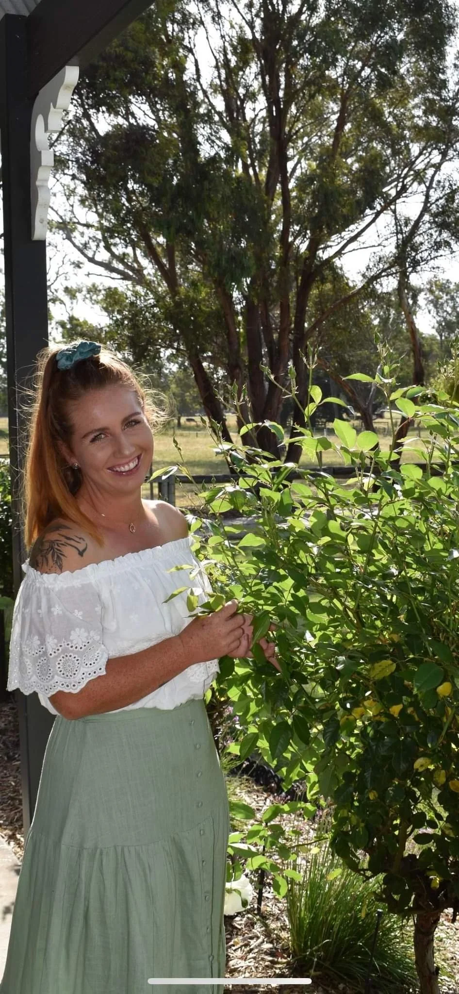 A woman with red hair, wearing a white off-shoulder top and a light green skirt, standing outdoors and smiling while holding green leaves in front of a large tree and bright sky.