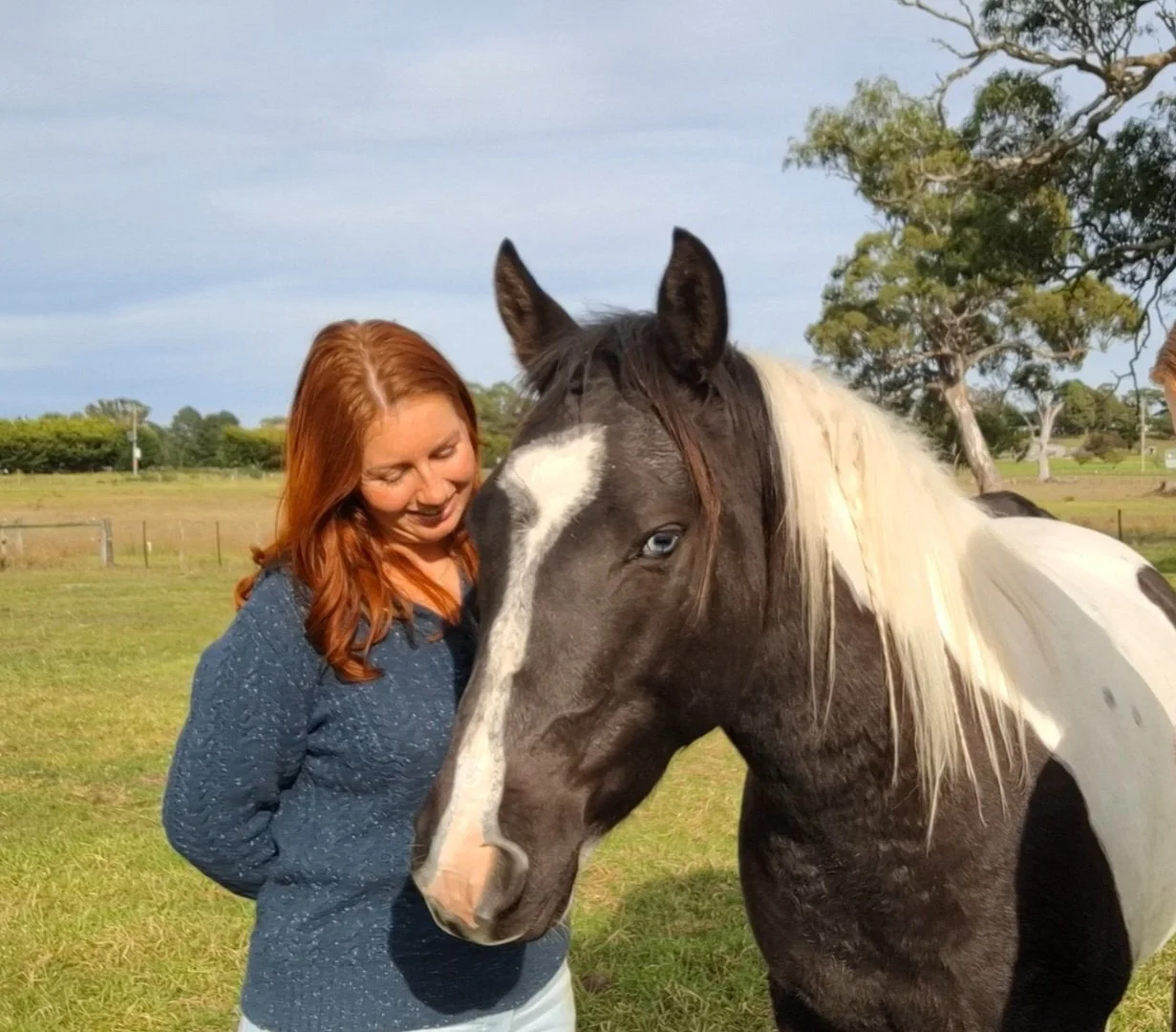 A woman with red hair smiling while petting a black and white horse in a grassy field.
