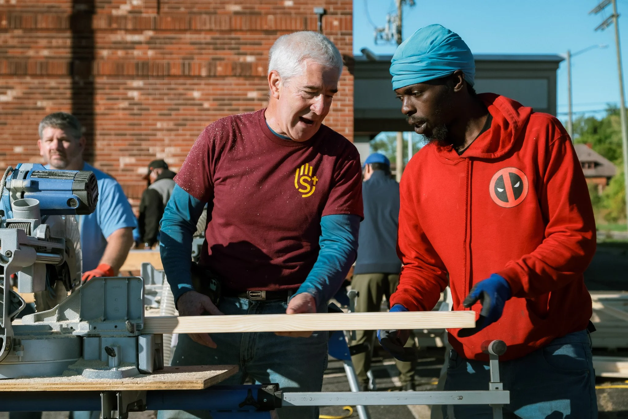 Two men working with a piece of wood at an outdoor construction site, with a third man watching in the background.
