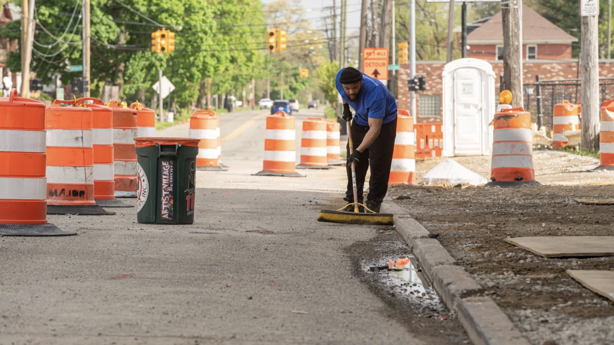 Man working on street construction, using a broom to sweep the road near orange and white traffic barrels and cones.