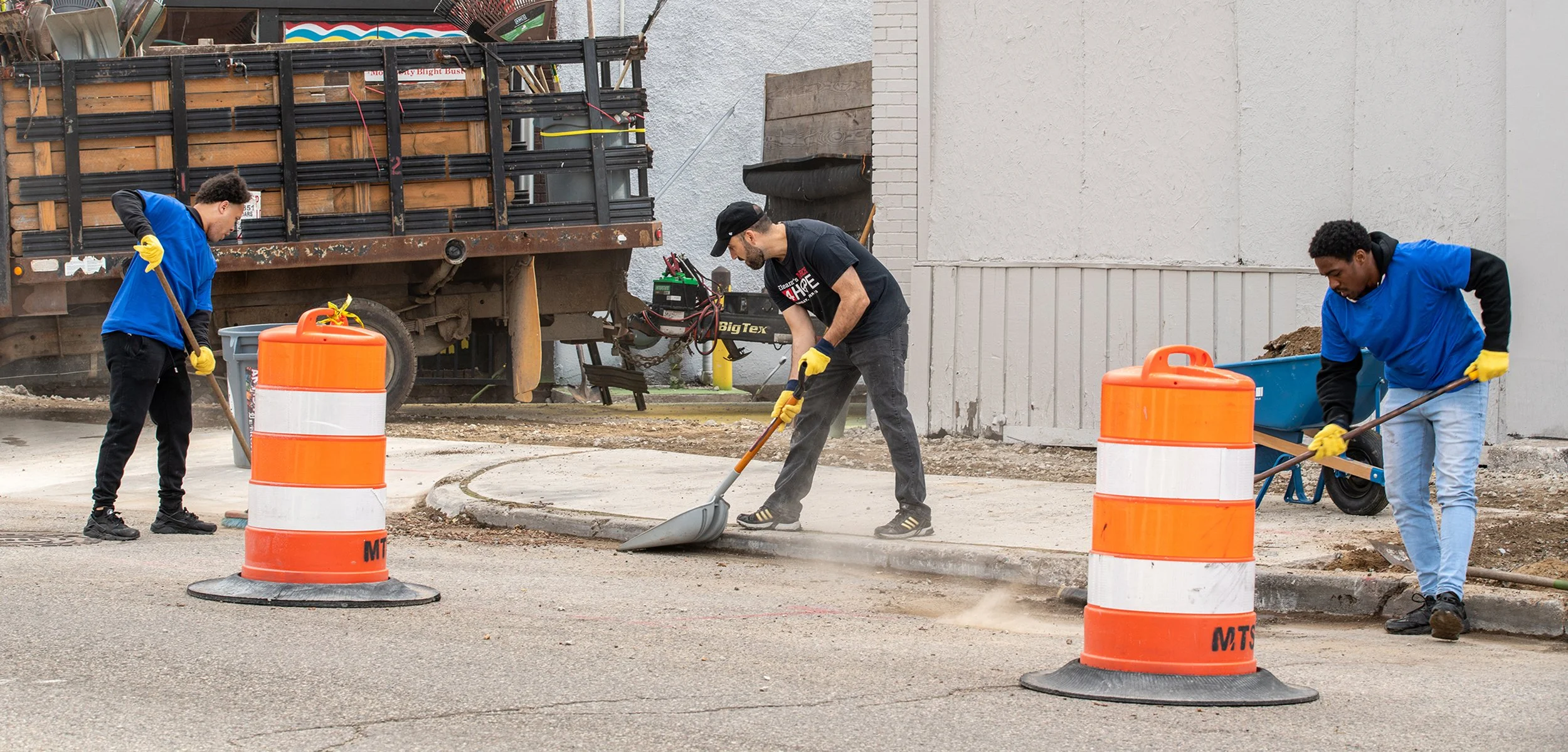 Three workers in blue shirts and yellow gloves working on a street repair project. Two of them are shoveling dirt or gravel, and the third is using a hand tool to smooth out the asphalt. Orange traffic barrels are positioned on the street to block the area, and a blue wheelbarrow is next to one worker. A truck and construction equipment are in the background.