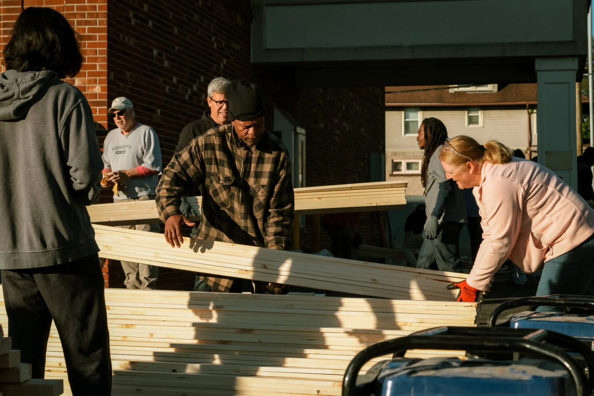 Group of people working together outdoors, handling wooden planks, likely construction or community project, under sunlight near a brick building.