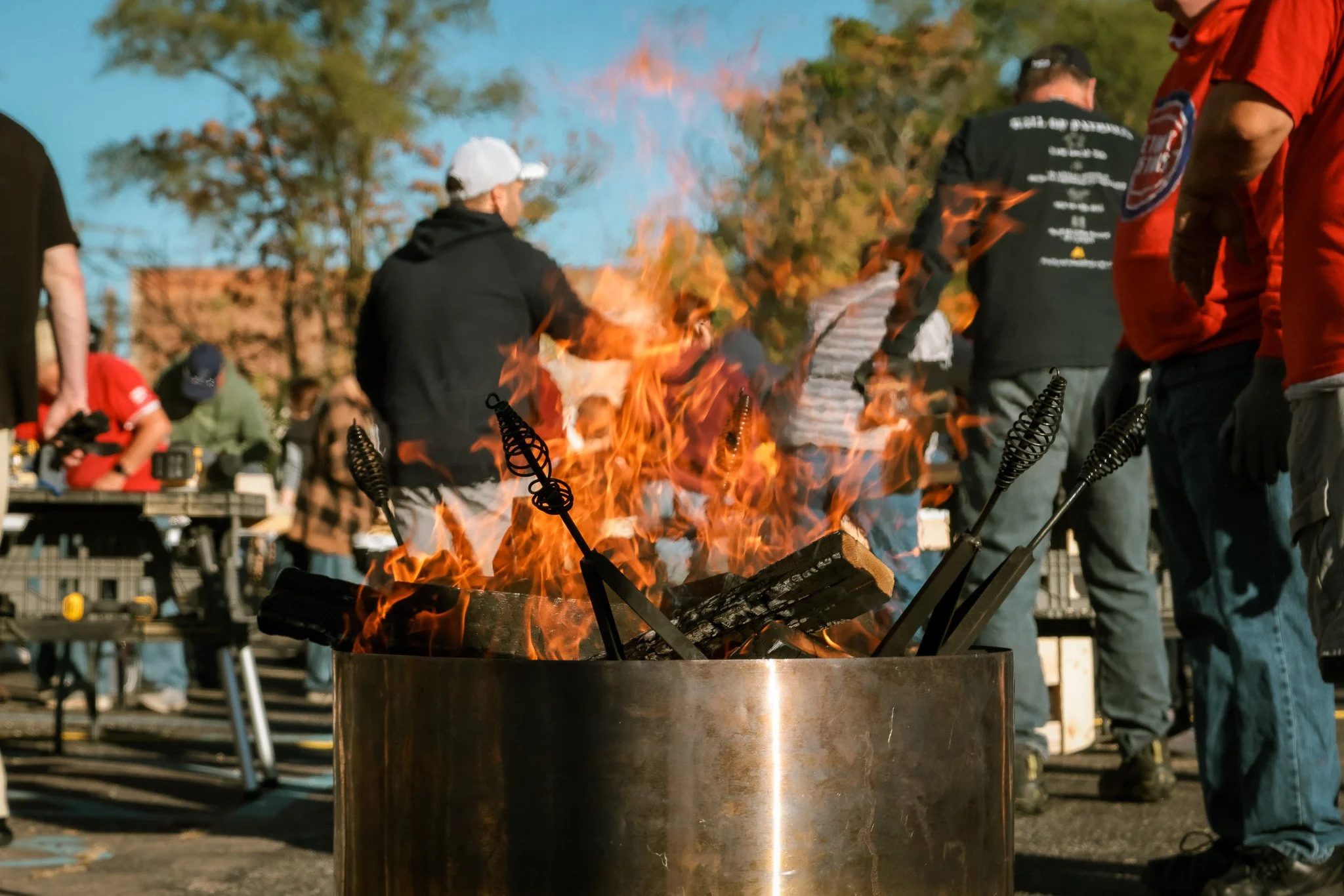 People gathered around an outdoor fire pit with logs and roasting sticks, on a clear day with trees in the background.