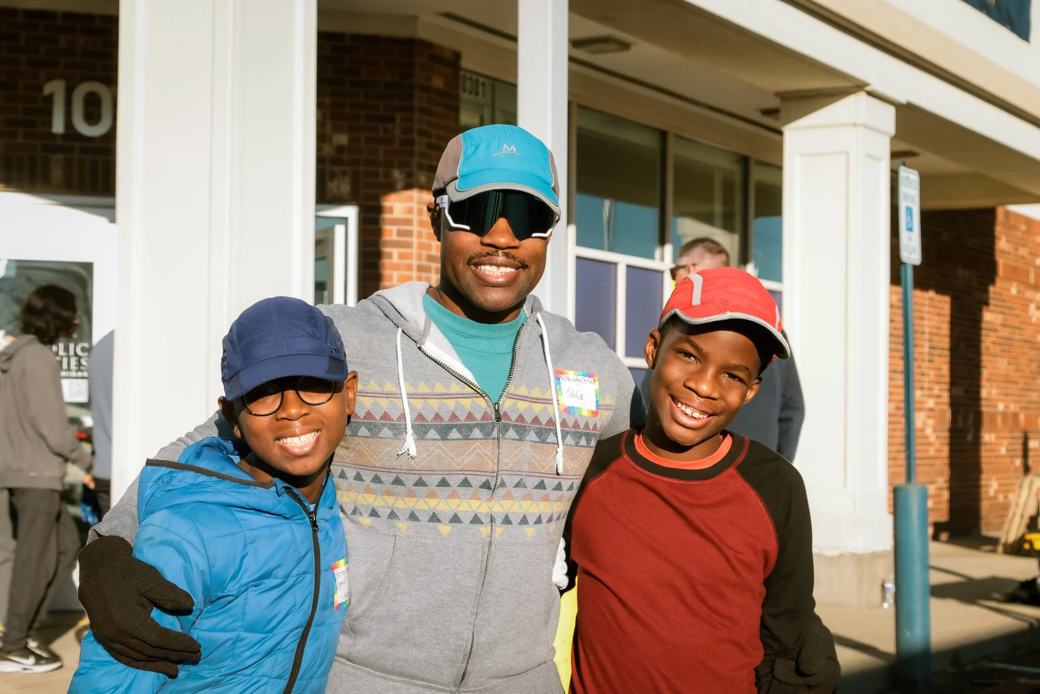 Three smiling males standing outdoors, one adult male with a beard, wearing sunglasses, a blue and gray cap, a gray patterned hoodie, and a teal shirt. Two young boys, one in a blue jacket and glasses with a blue cap, and the other in a red and black