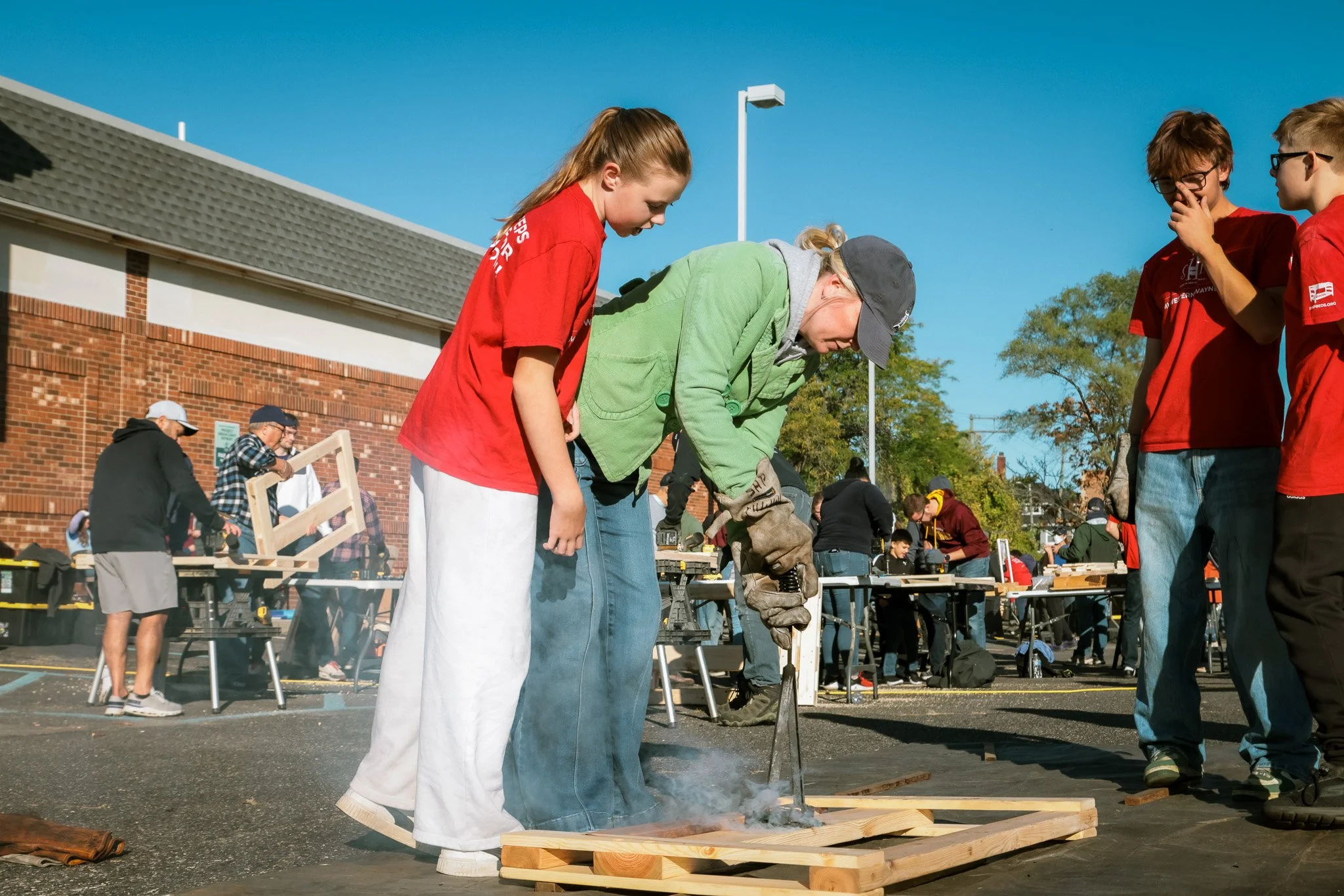 Group of people working on a woodworking project outdoors, with a woman cutting wood and others observing, under a bright blue sky.
