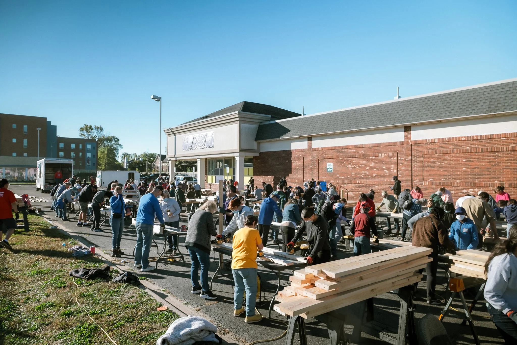 Large group of people working together outdoors at a long assembly line with pieces of wood, outside a commercial building on a sunny day.