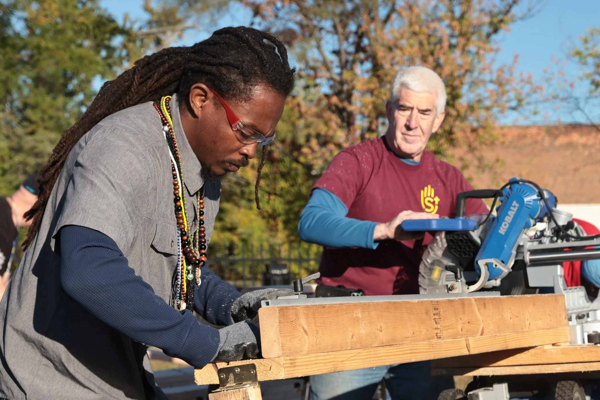 Two men working on a wooden structure outdoors in a park with trees and clear sky, one using a miter saw, the other helping with the project.