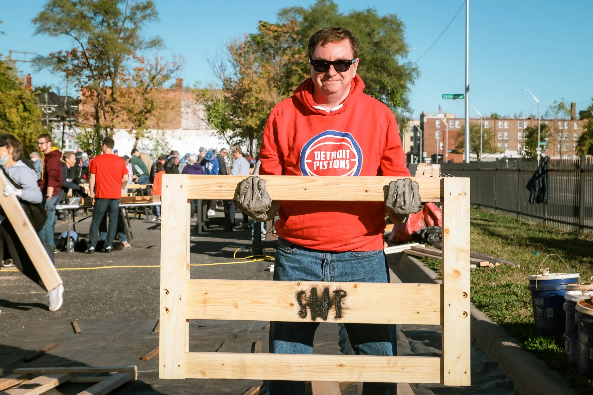 A young man wearing sunglasses and a red Detroit Pistons hoodie holds a wooden pallet with the word 'SLUT' spray-painted on it, during an outdoor community event with many people working together in the background.