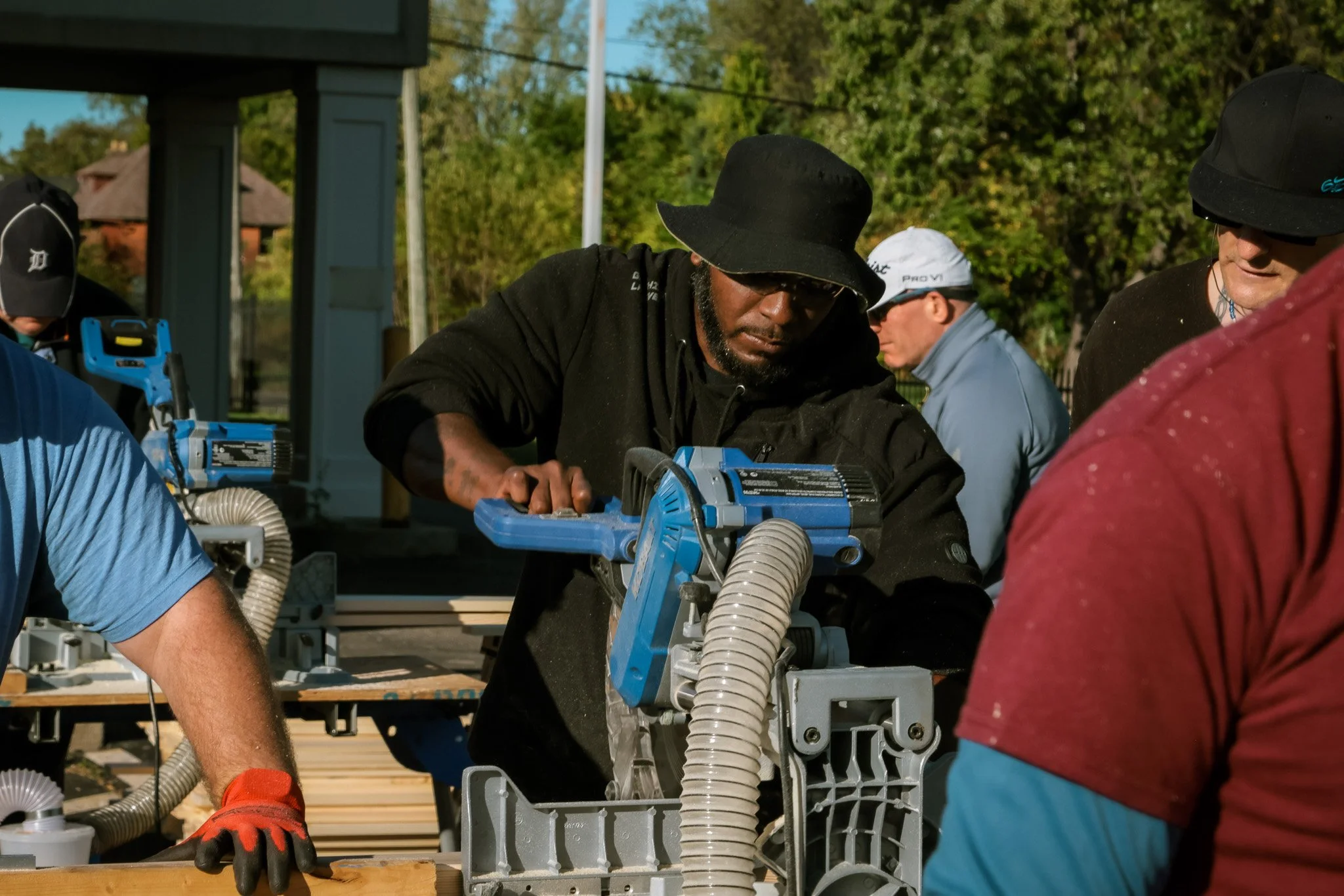 Group of people working outdoors using a power saw, with trees and a building in the background.