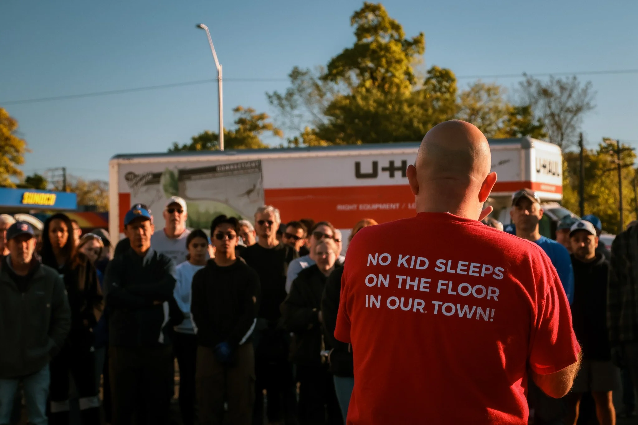 A man in a red shirt with text on the back speaking to a crowd at an outdoor event, with trees and trucks in the background.