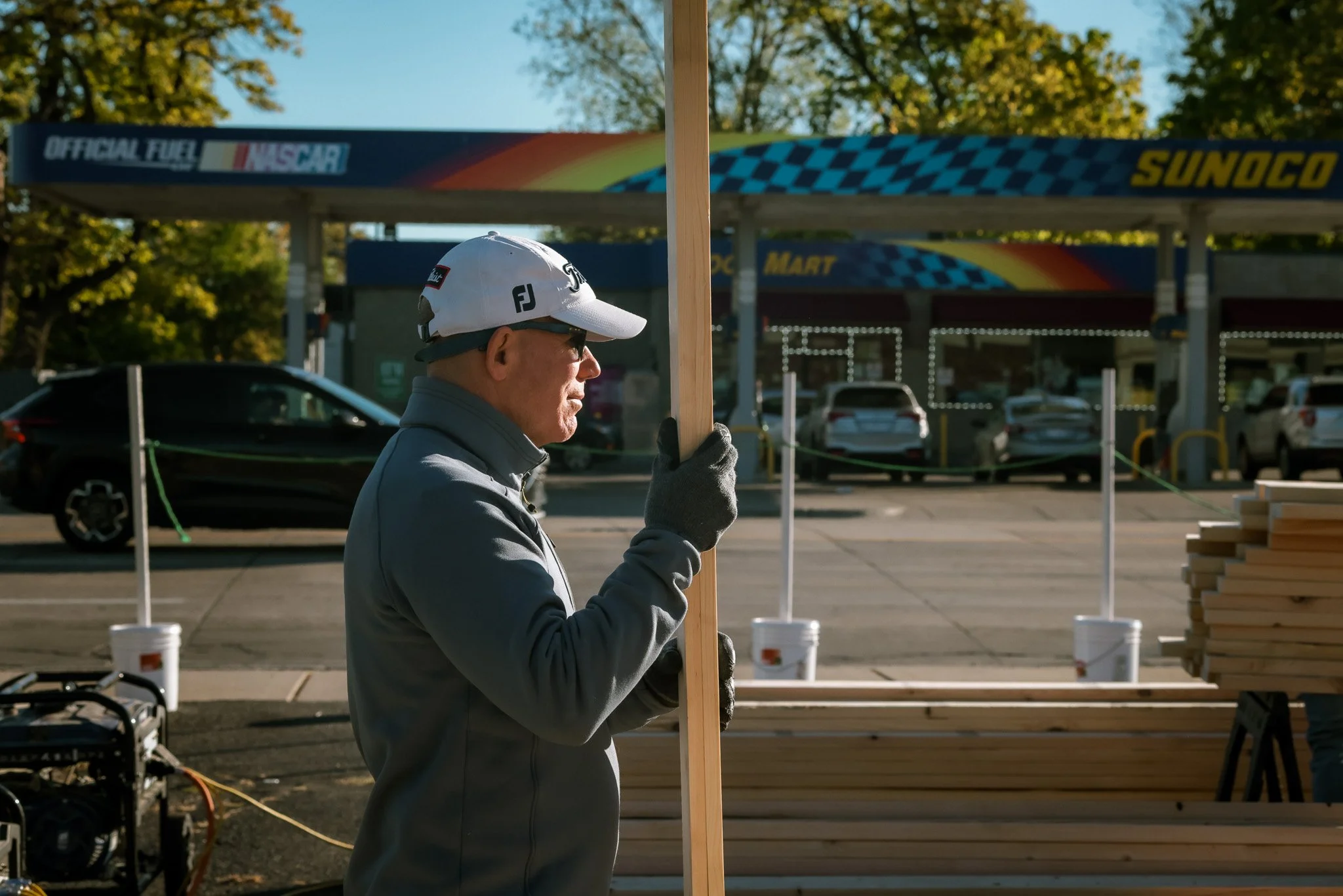 A man in a gray jacket, black gloves, and a white baseball cap holding a wooden board at an outdoor construction site. There are parked cars and a Sunoco gas station in the background.