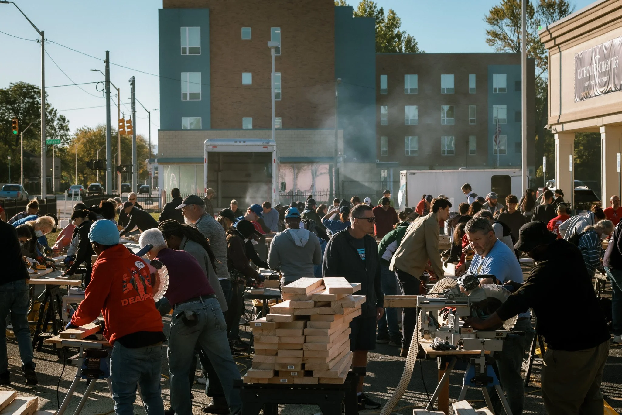 A large group of people working with woodworking tools outdoors in front of a building with a bright sky and trees in the background.