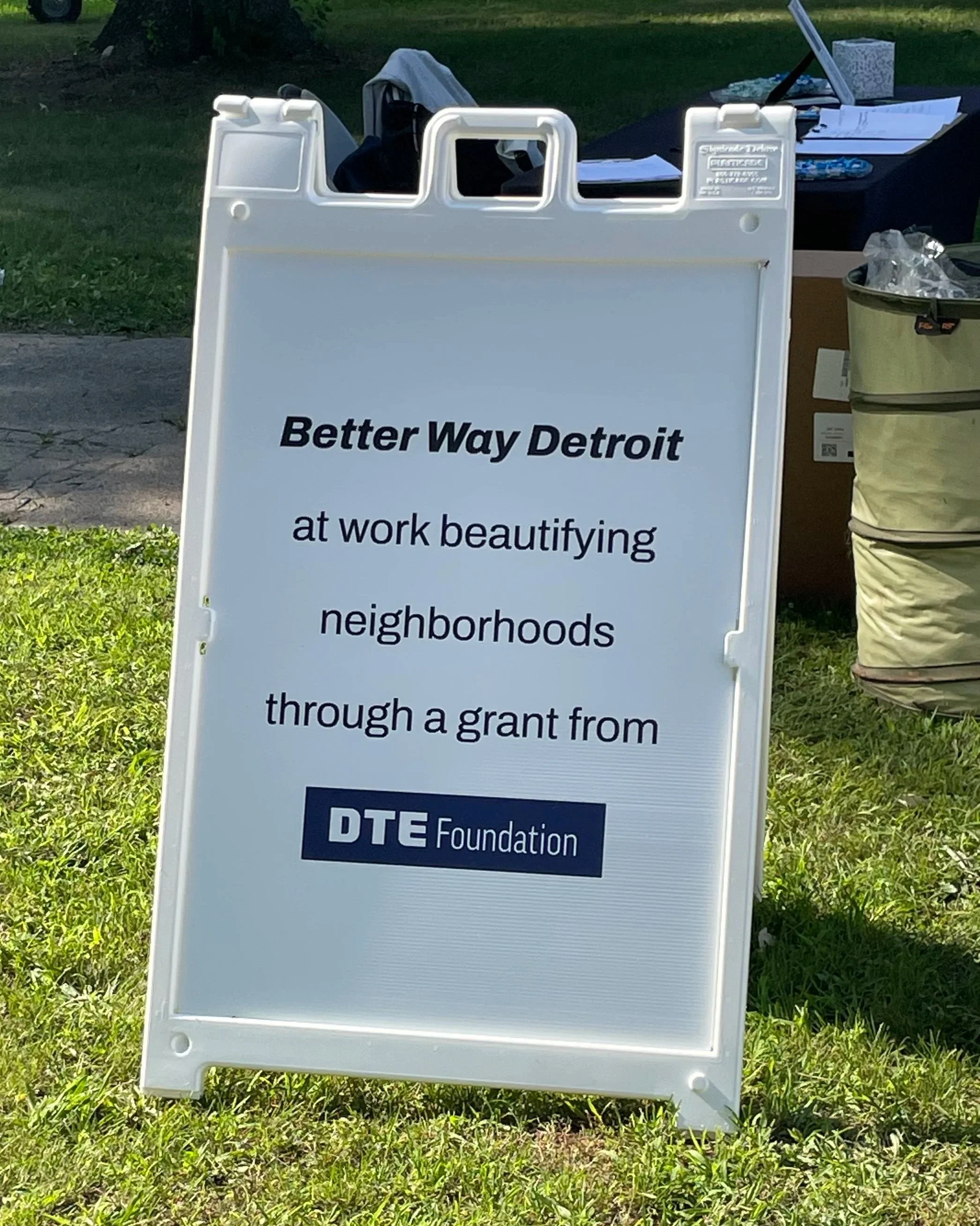 A white roadside sign promoting the Better Way Detroit organization, describing their work beautifying neighborhoods through a grant from the DTE Foundation, with some tables and bushes in the background.