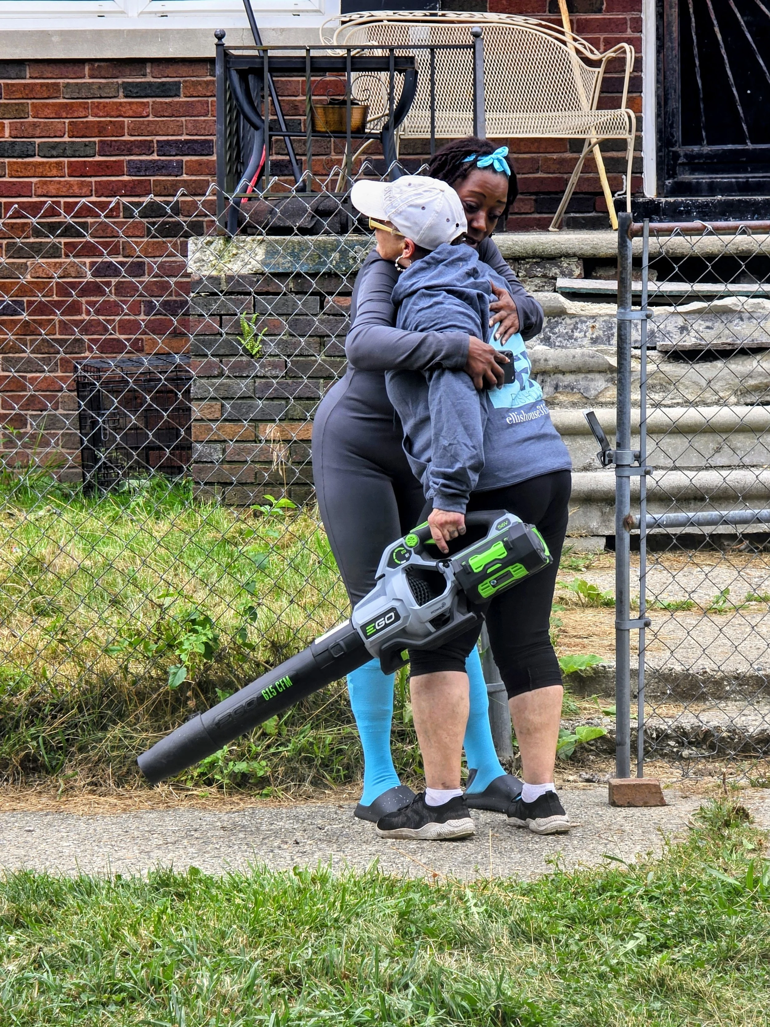A woman hugging a young girl outdoors, the woman holding a leaf blower, with a chain-link fence, brick steps, and a house in the background.