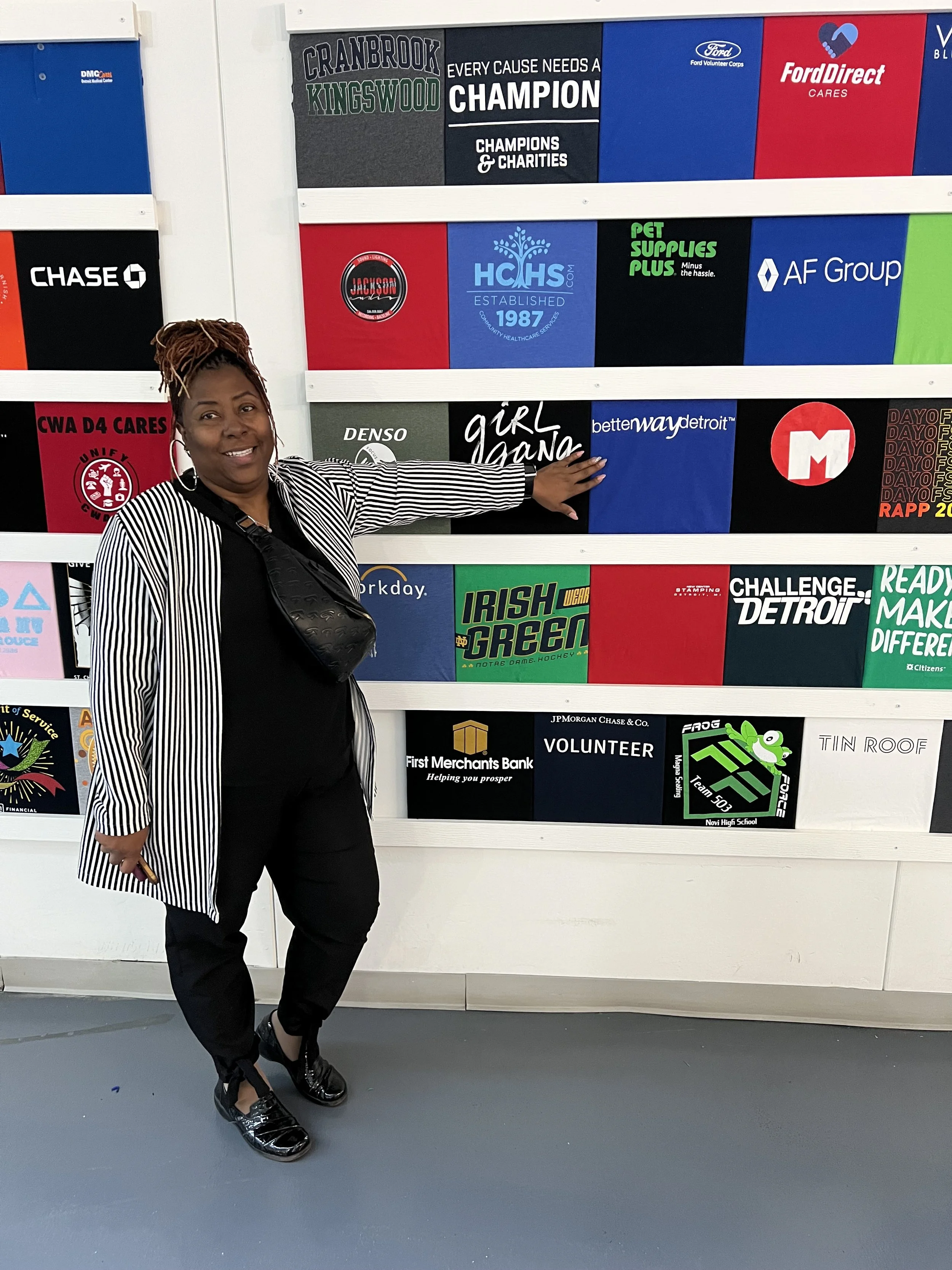 A woman with short dreadlocks and hoop earrings smiling and pointing at a display of various colorful sponsor logos and promotional signs on a wall.