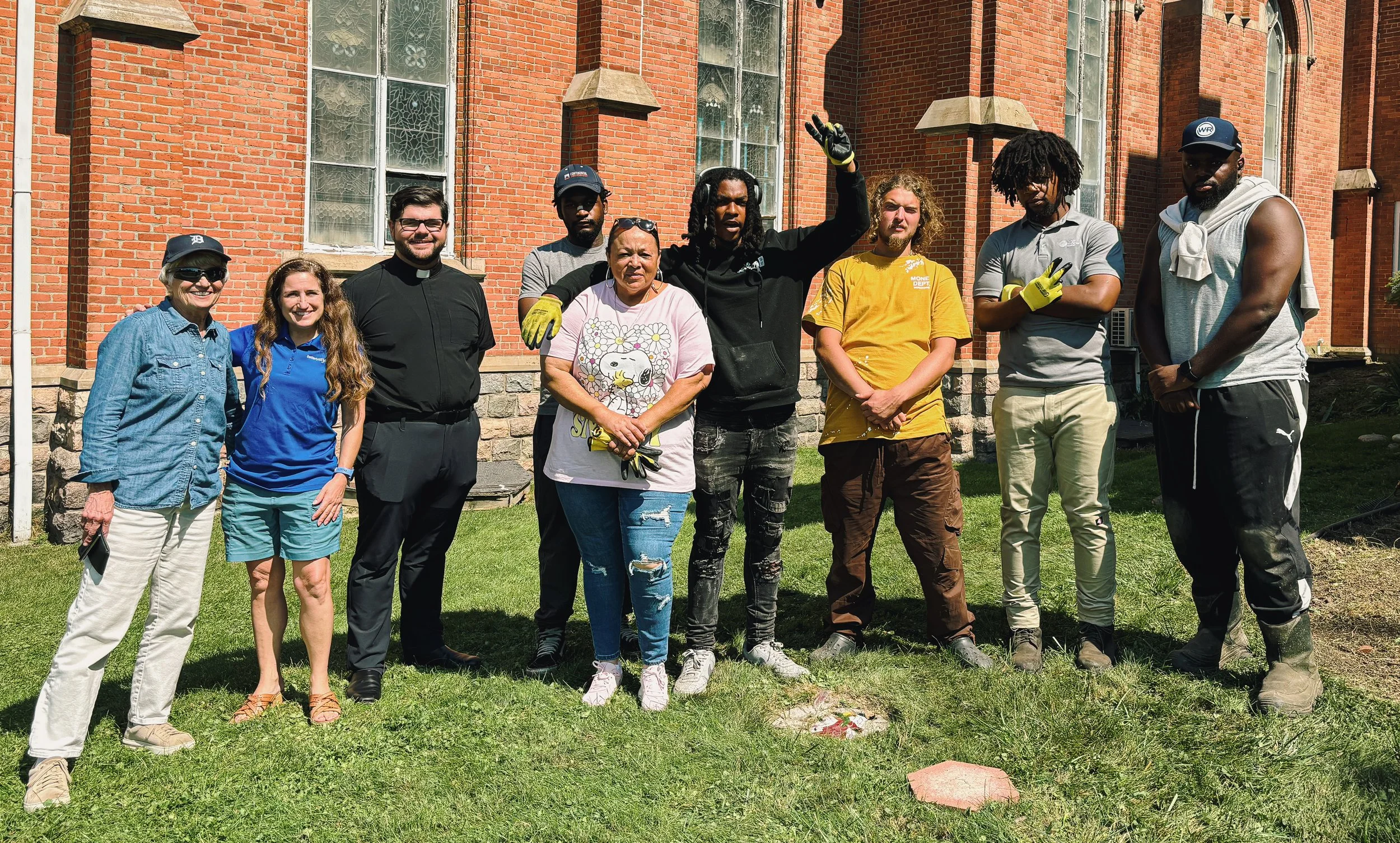 A group of nine diverse people standing on a grassy area in front of a red brick building with large windows, some smiling and posing for the photo.