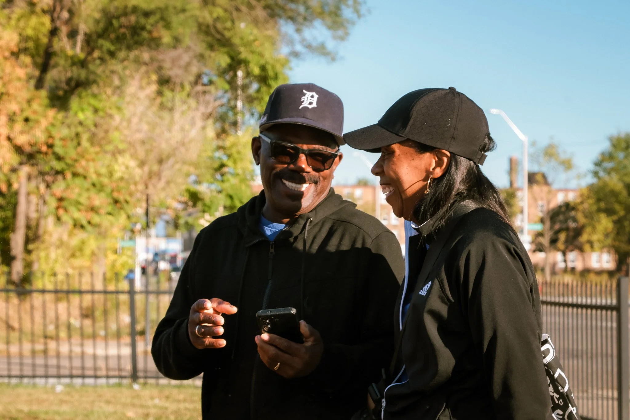 Two smiling people in black jackets and caps looking at a phone outdoors on a sunny day, with trees and a fence in the background.