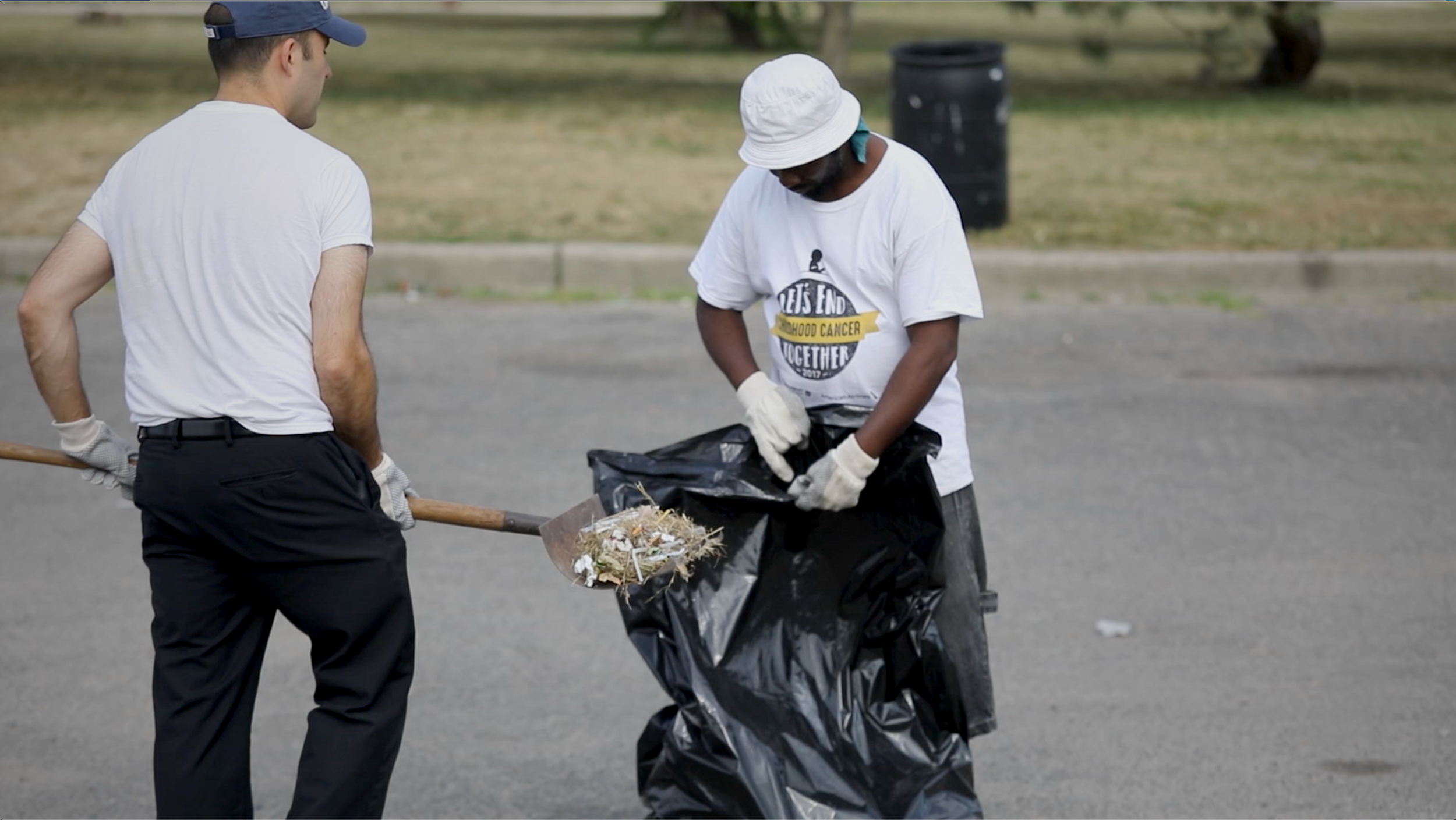 Two men participating in a community cleanup; one man is shoveling trash into a black garbage bag while the other stands nearby with a trash picker, both wearing casual clothes and gloves.