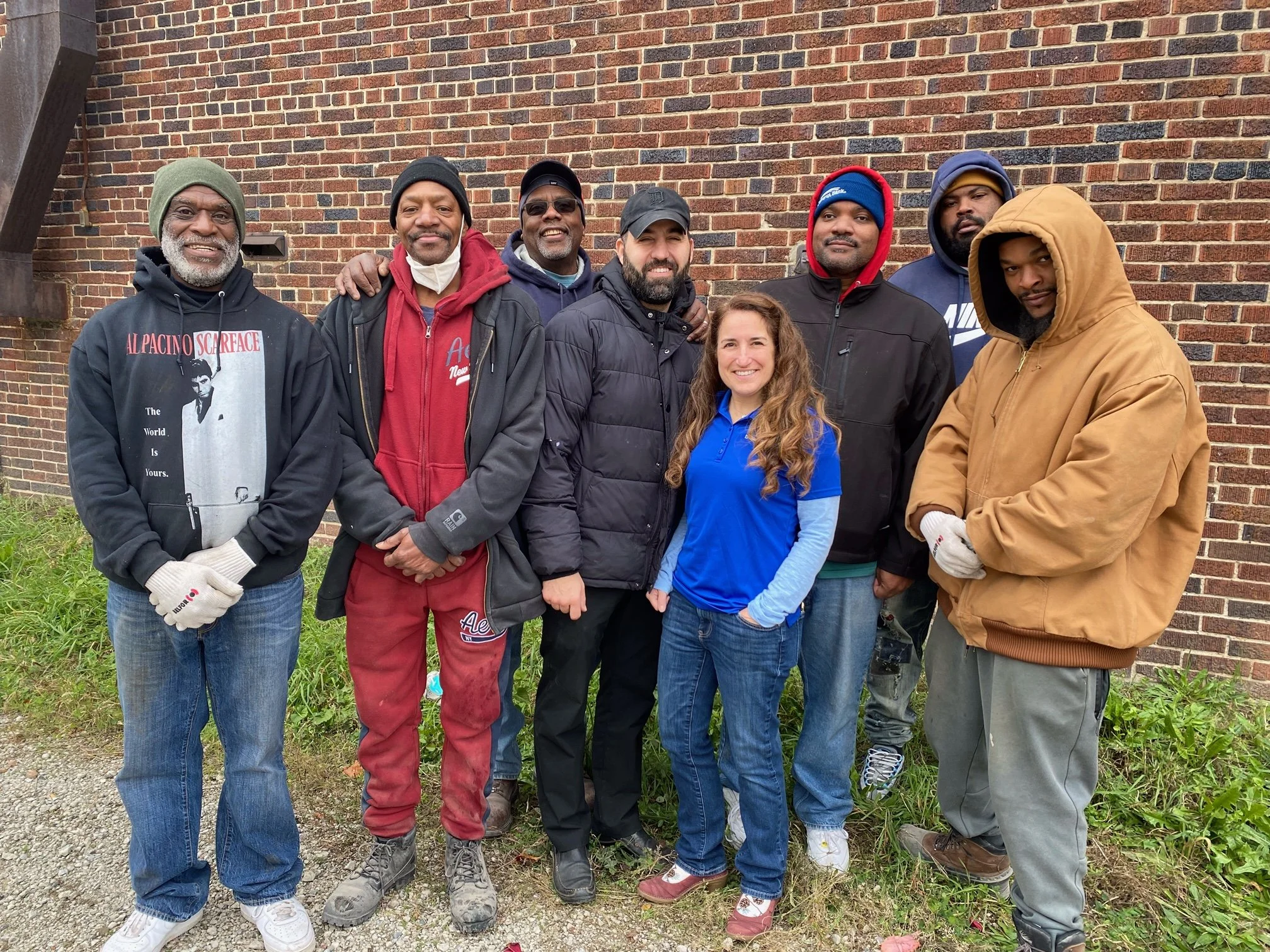 A diverse group of nine people standing outdoors in front of a brick wall, dressed in casual and warm clothing.