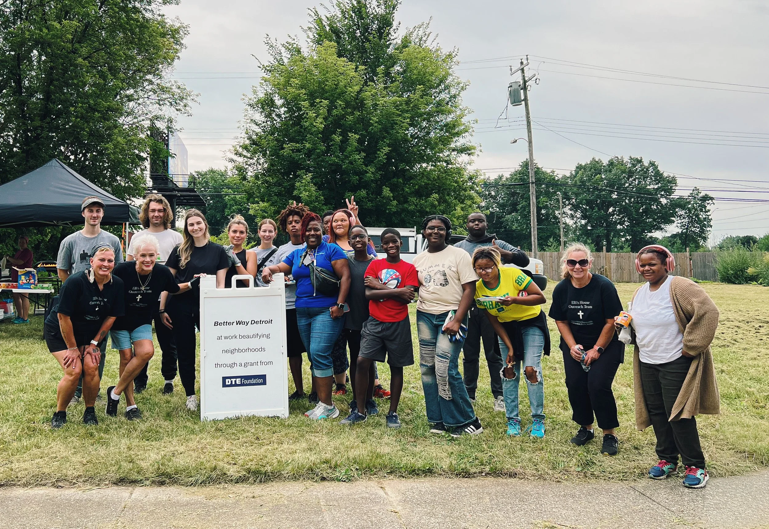 Group of diverse people posing outdoors in a park-like setting, some smiling and making peace signs, with a sign that reads 'Better Way Detroit at work beautifying neighborhoods through a grant from DTE Foundation.'