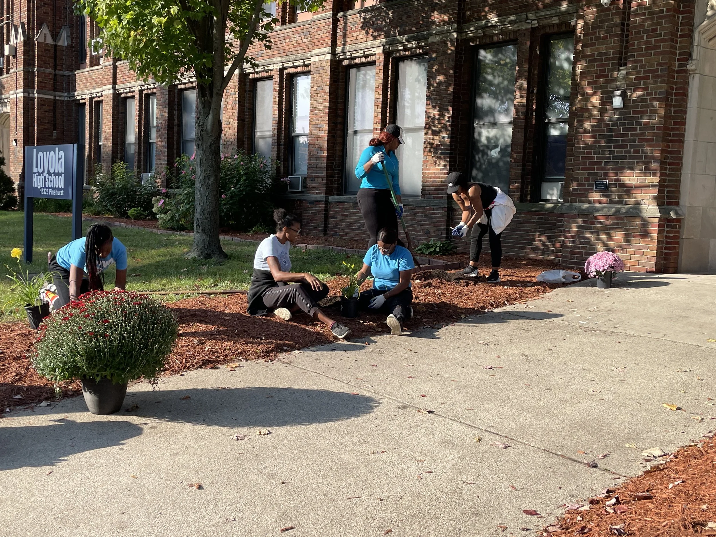 Group of people planting flowers in a flower bed outside of Loyola High School on Pinehurst street.