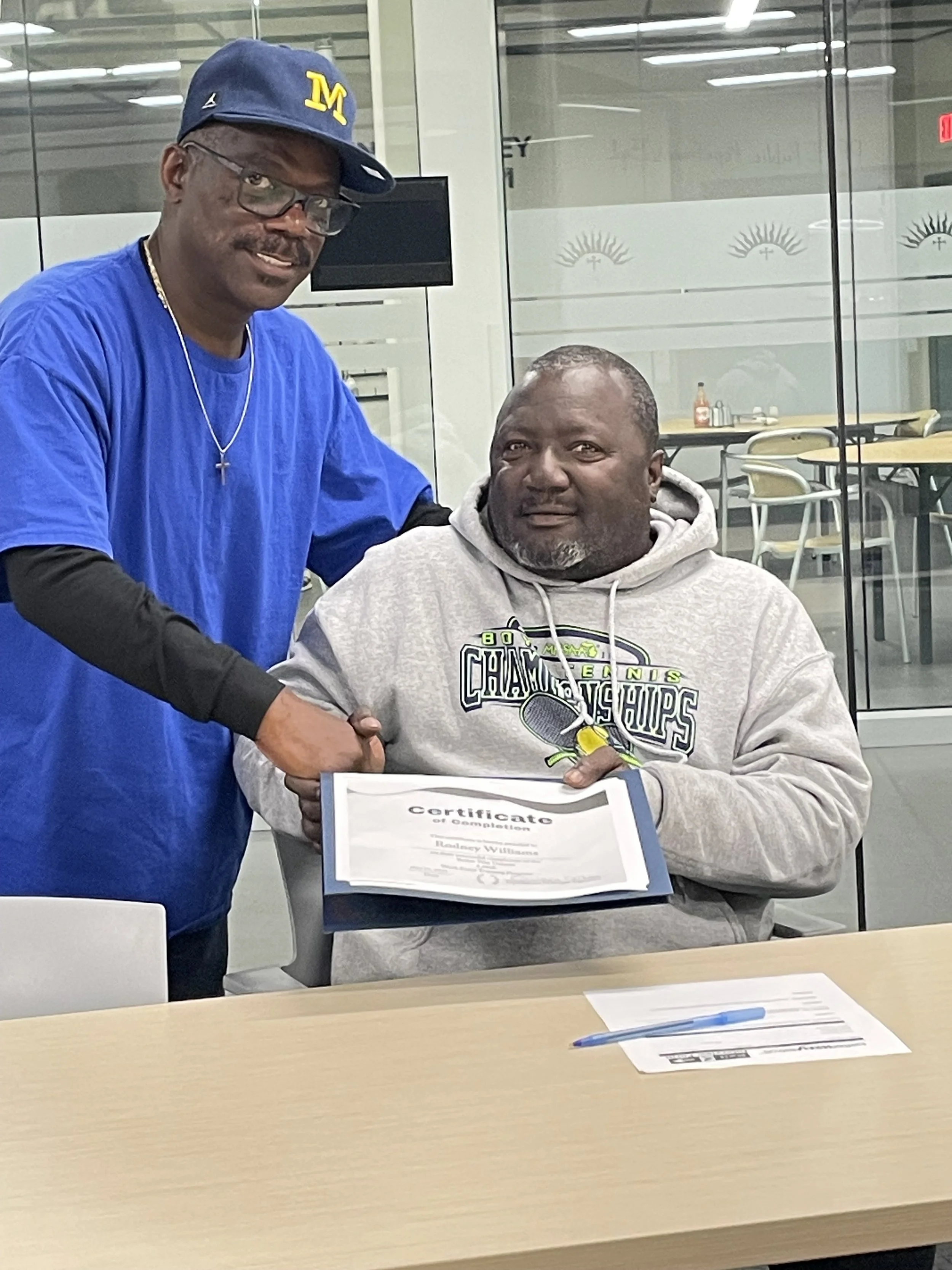 Two men are in an indoor setting, one standing and one sitting. The man standing is wearing a blue T-shirt, glasses, a navy cap with a yellow 'M' on it, and a silver necklace. The man sitting is holding a certificate and is wearing a gray hoodie with a tennis or sports team logo. They are shaking hands, and there is a table with a paper and pen in front of them.