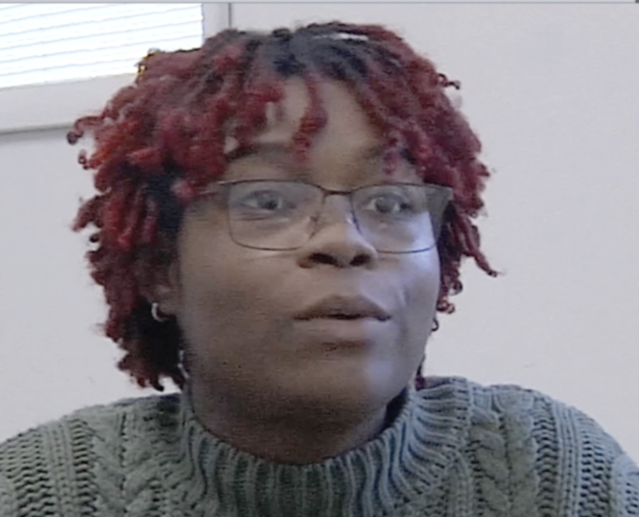 A person with short, curly, red dreadlocks wearing glasses and a light-colored, patterned sweater, looking slightly to the side indoors.