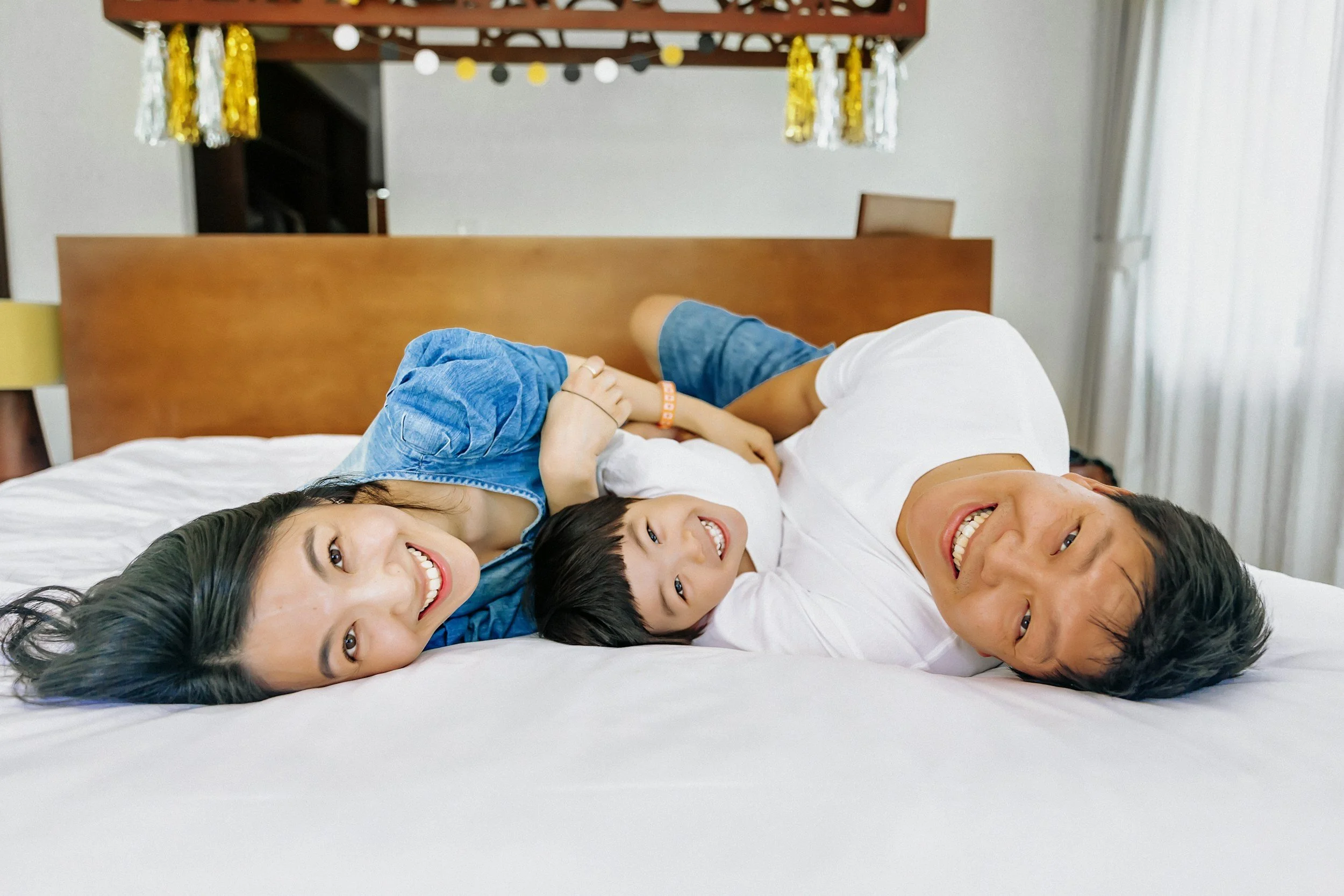 A family lying on a bed, smiling and taking a selfie together. The family includes two women and two young boys. The background shows a bed headboard and decorated hanging garlands.