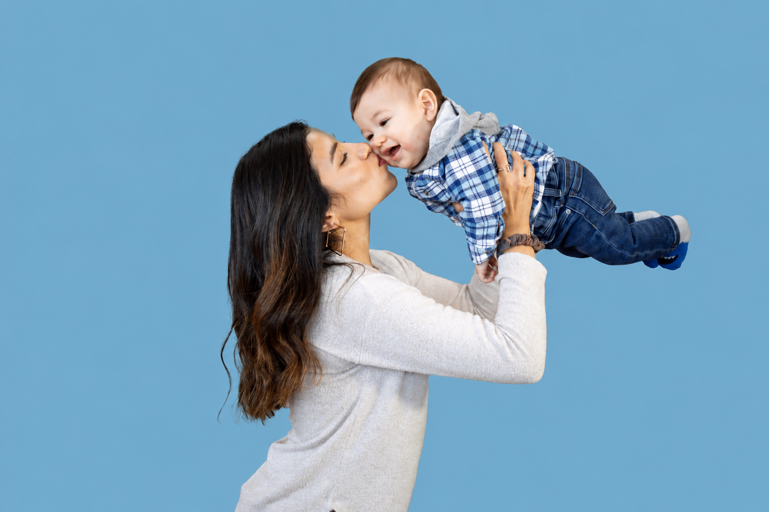 A woman holding a young boy up in the air while touching their noses together against a light blue background.