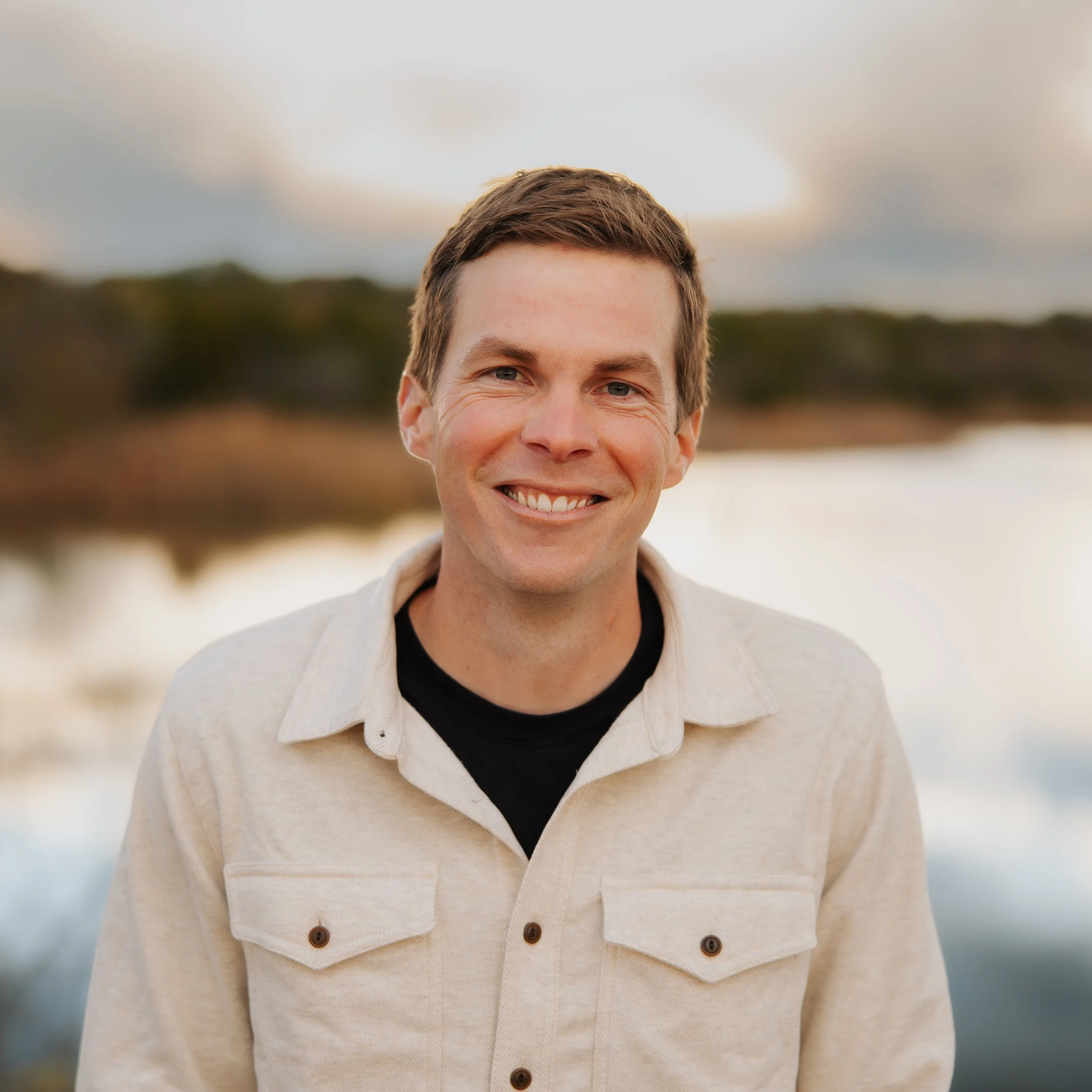 Close-up of a smiling man wearing a cream-colored jacket with brown buttons over a black shirt, outdoors during daytime, with blurred natural background.