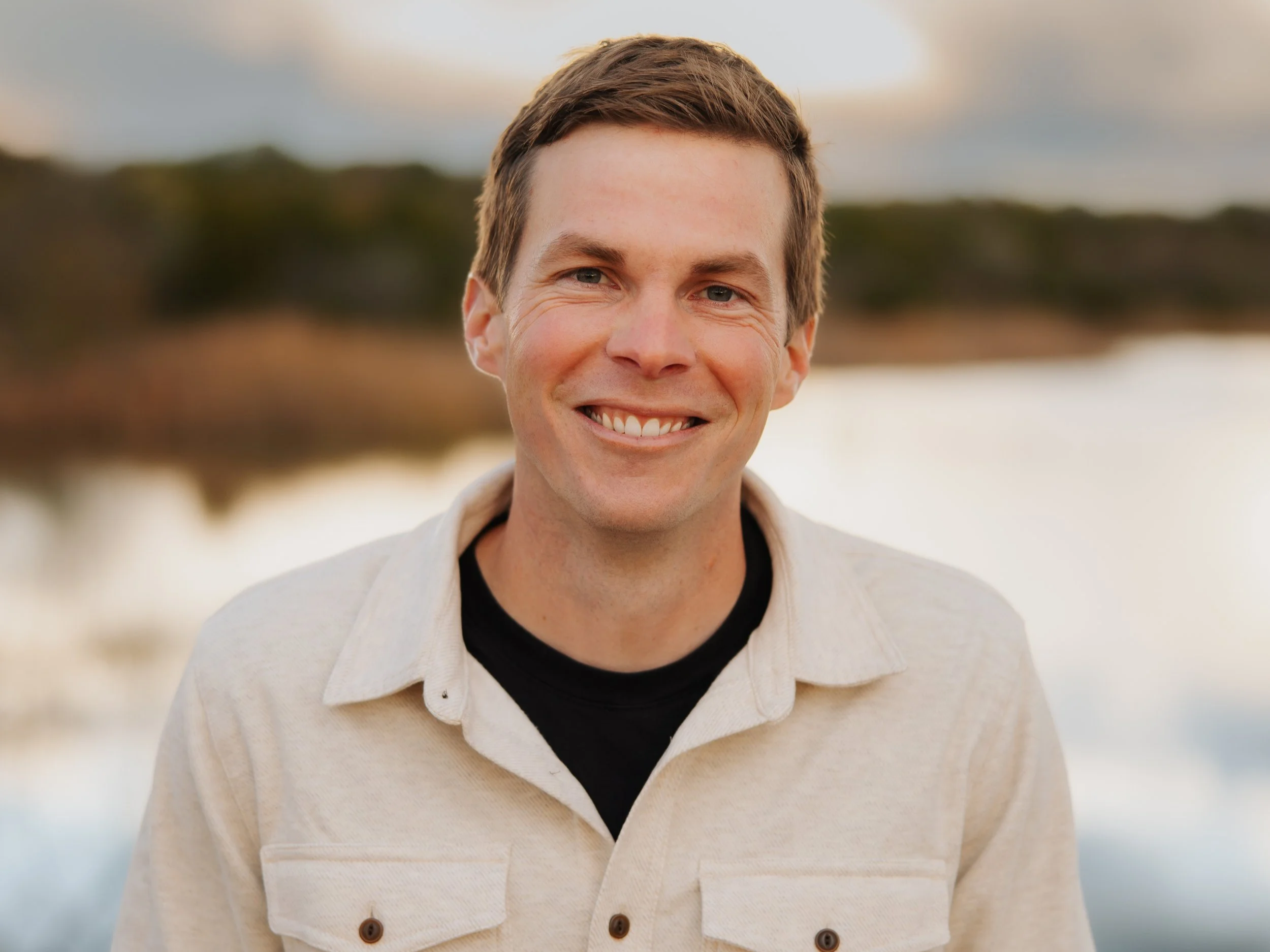 A close-up of a smiling man outdoors with blurred natural background, wearing a beige jacket over a black shirt.