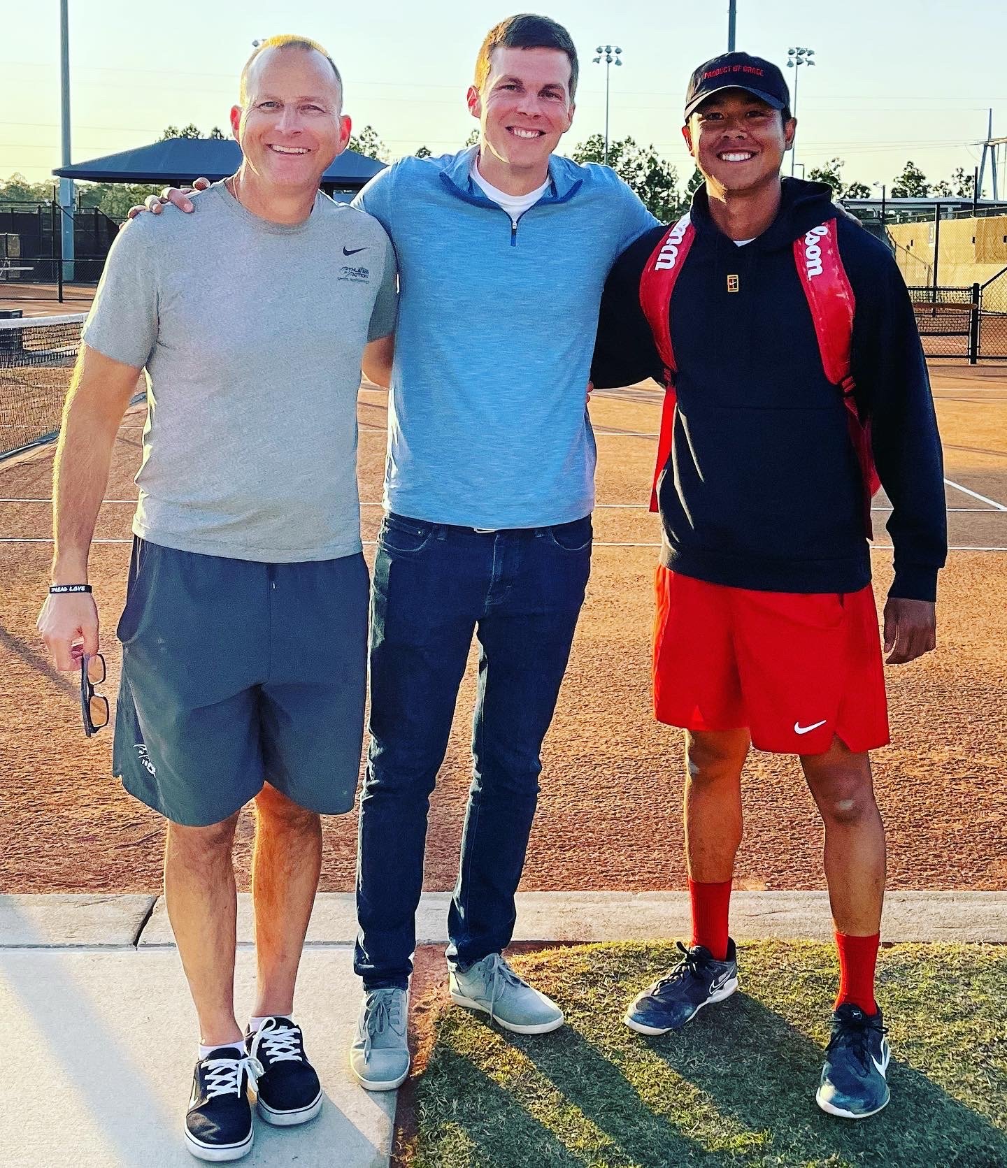Three men standing on a tennis court, smiling at the camera. The man on the left is wearing a gray Nike t-shirt, gray shorts, and black-and-white sneakers. The man in the middle is wearing a blue zip-up jacket, jeans, and white sneakers. The man on the right is wearing a black hoodie, red shorts, a black cap, and red socks, with a red tennis bag on his back.