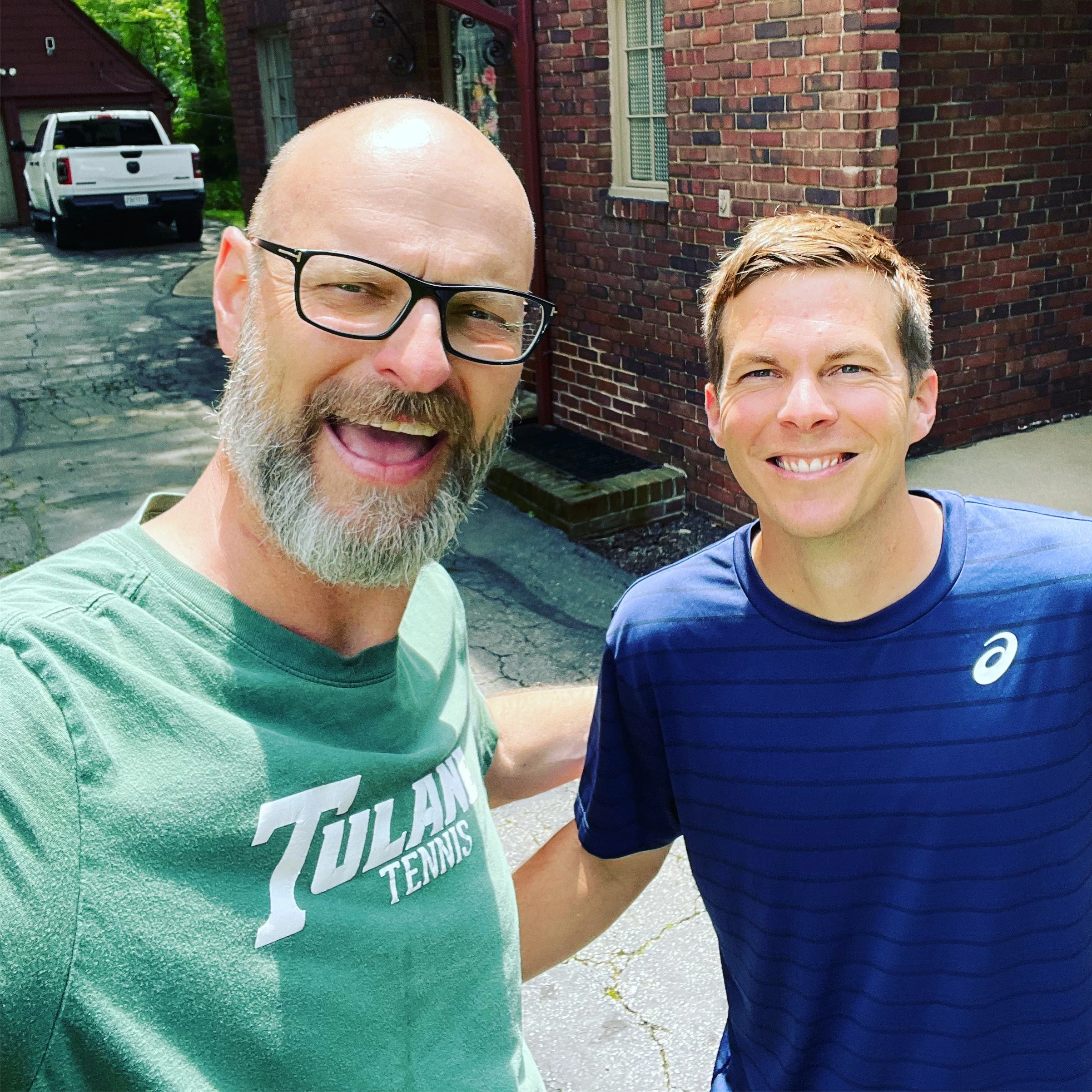Close-up of two smiling men, one with glasses and a beard wearing a green shirt, and the other with short hair in a blue shirt, standing outside in front of a brick house with a parked white pickup truck.