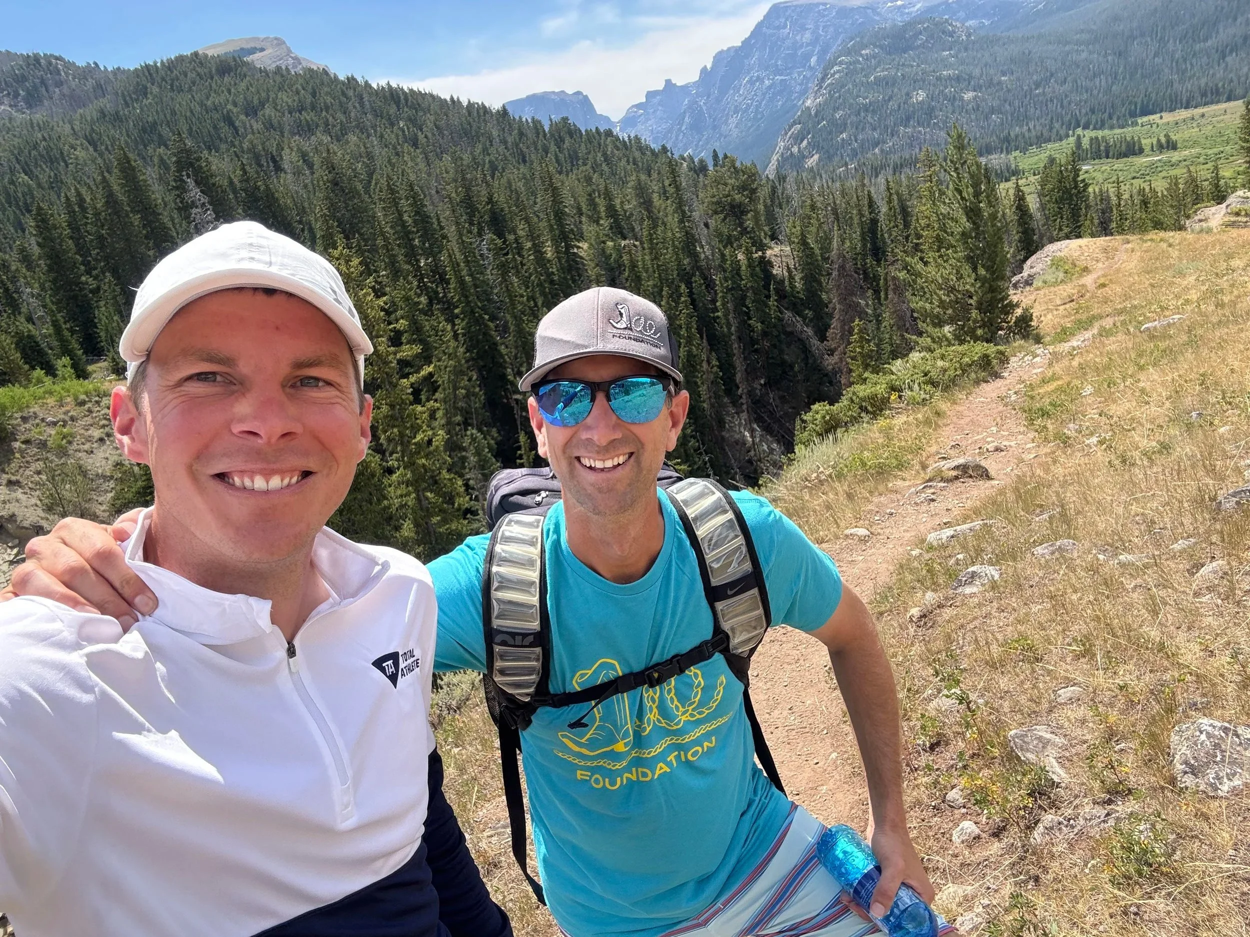 Two men hiking in a mountainous forest, smiling, wearing athletic clothing and backpacks.