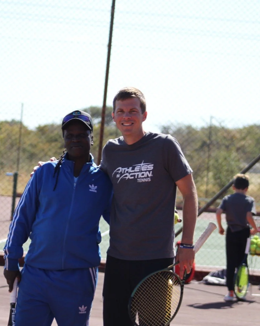Two men on a tennis court, one in a blue tracksuit and cap, the other in a gray T-shirt and holding a tennis racket, smiling at the camera with a fence and other players in the background.