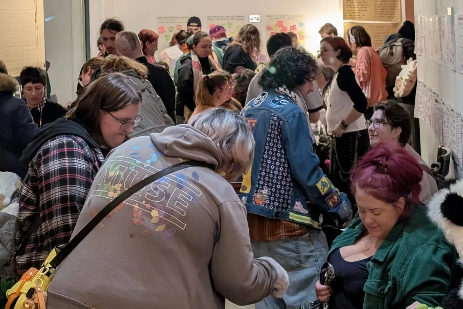 Group of people gathered in a crowded room, examining posters or charts on the walls.