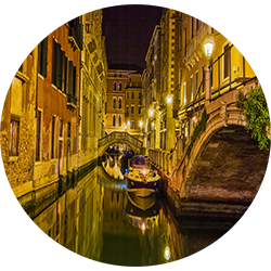 Nighttime view of a narrow canal in Venice, Italy, with a boat docked along the water and historic buildings illuminated by streetlights.