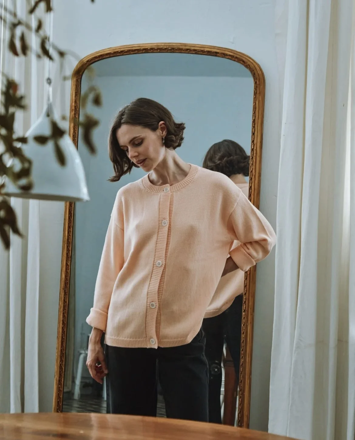 Une femme avec des cheveux court, portant un sweat rose pâle, se regarde dans un miroir en bois.