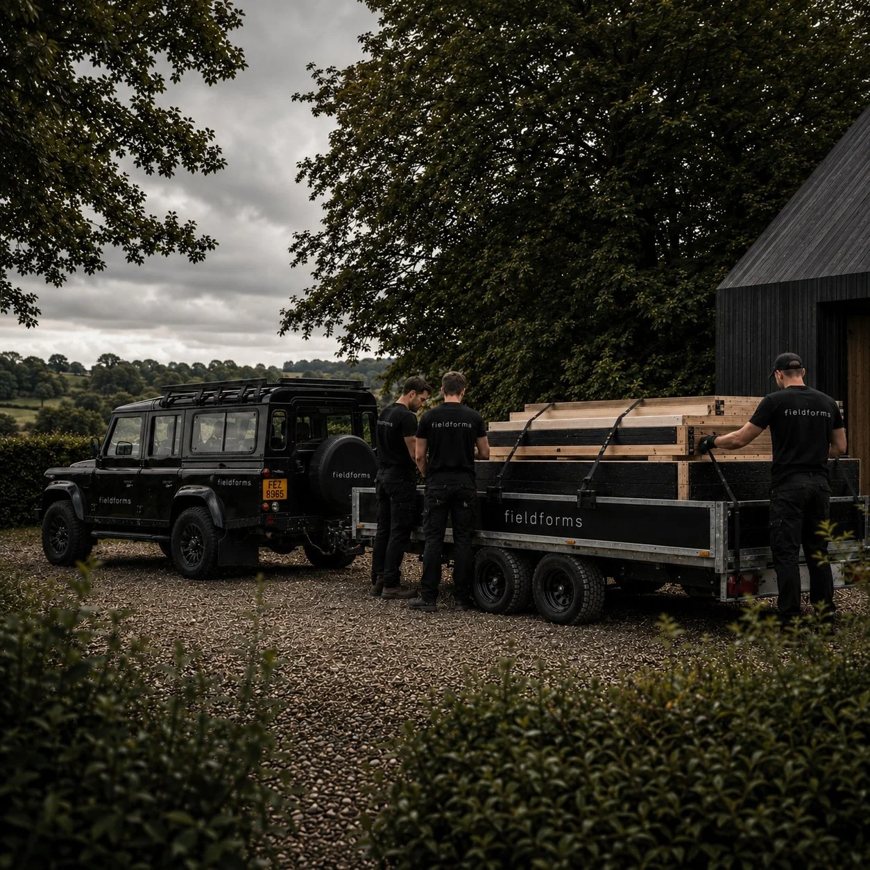Three men in black fieldforms shirts loading wooden beams onto a trailer attached to a black off-road vehicle, set in a rural landscape with trees and cloudy sky.