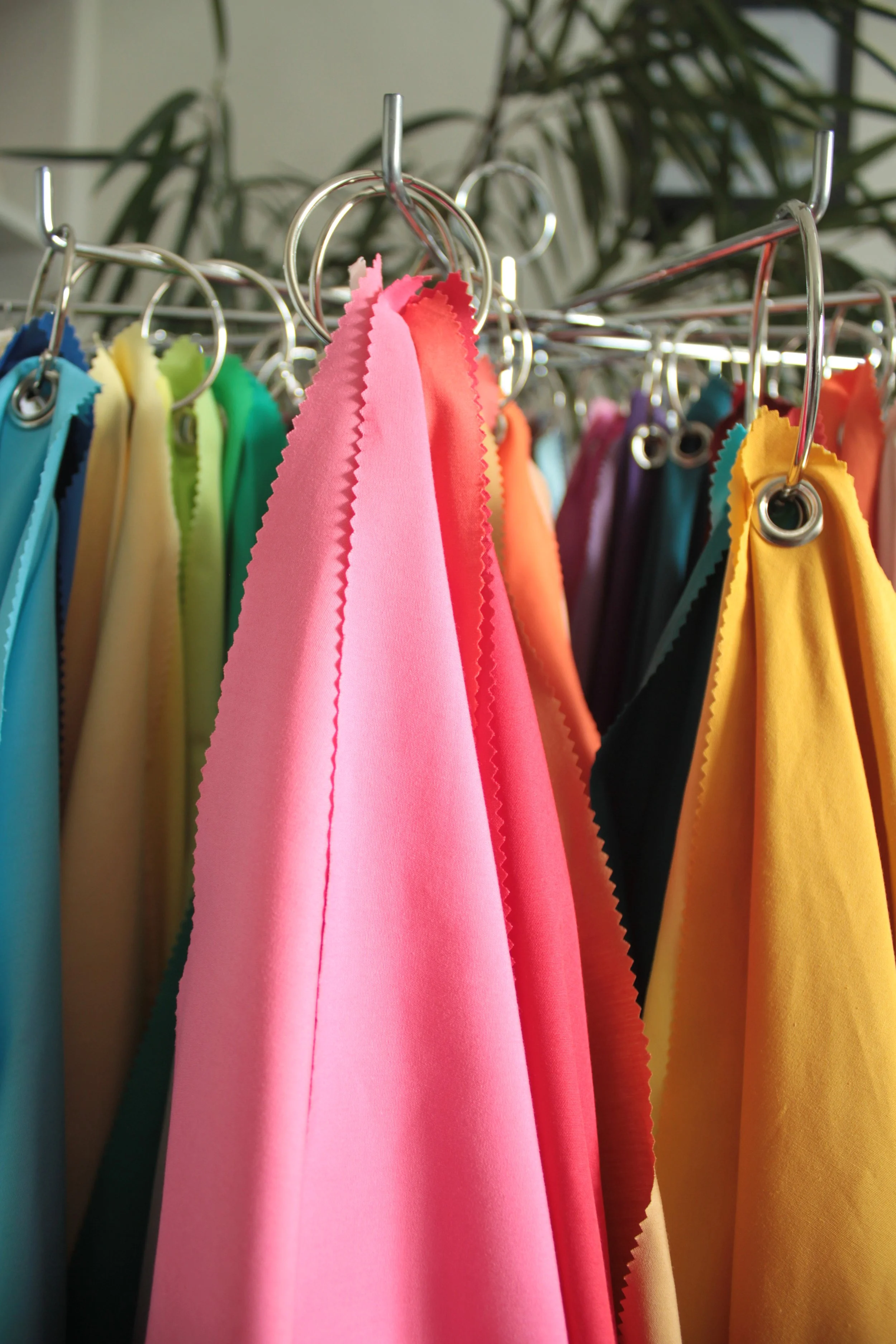 Colourful fabric drapes hanging on metallic hooks, arranged in a rainbow pattern with shades of blue, yellow, green, pink, purple, black, and gold, with a blurred background of indoor plants.